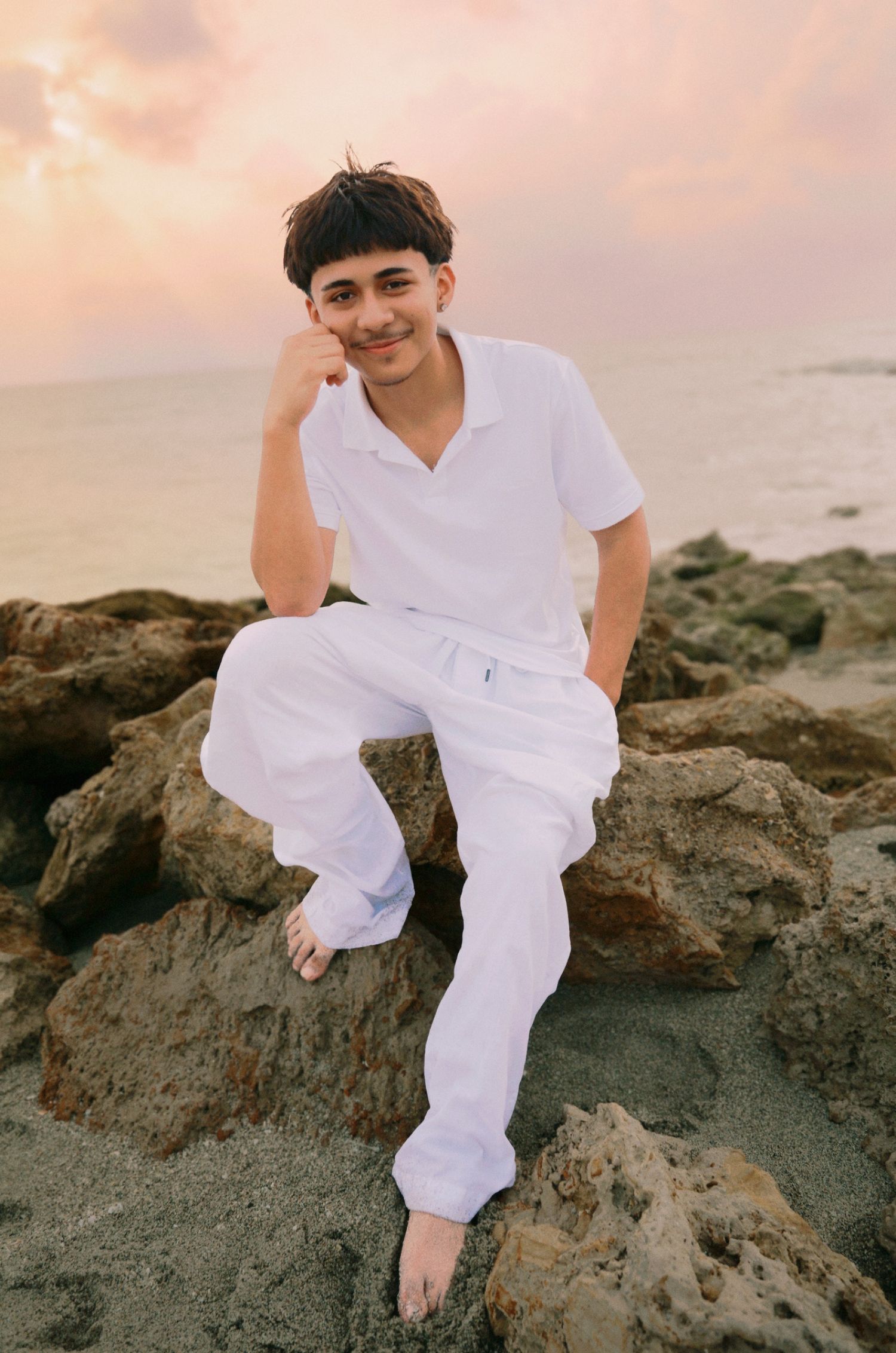 Young man in white outfit sitting on rocky beach at sunset, smiling confidently with ocean in background.