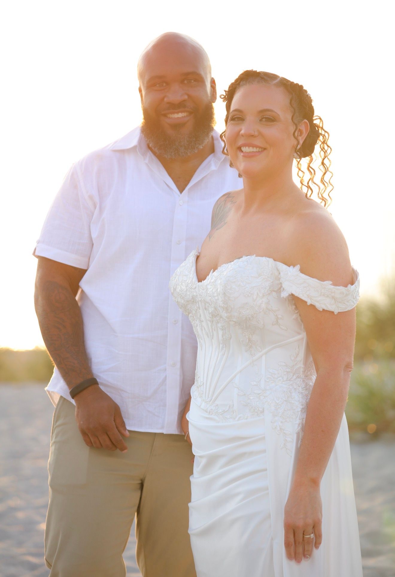 Couple in wedding attire smiling on beach at sunset, bride in white off-shoulder gown, groom in white shirt.
