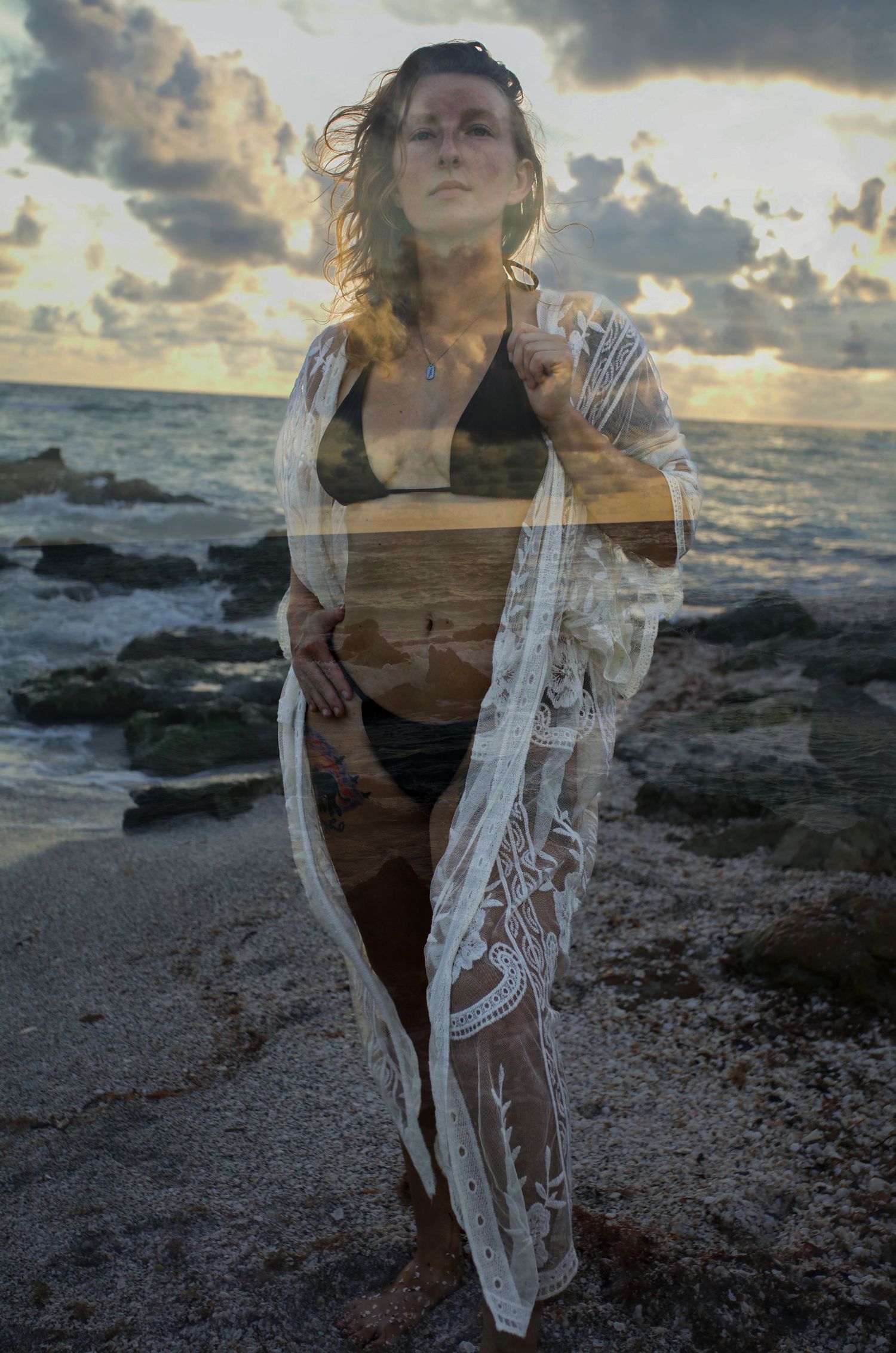 Woman in bikini and lace cover-up poses on rocky beach at sunset with dramatic cloudy sky.