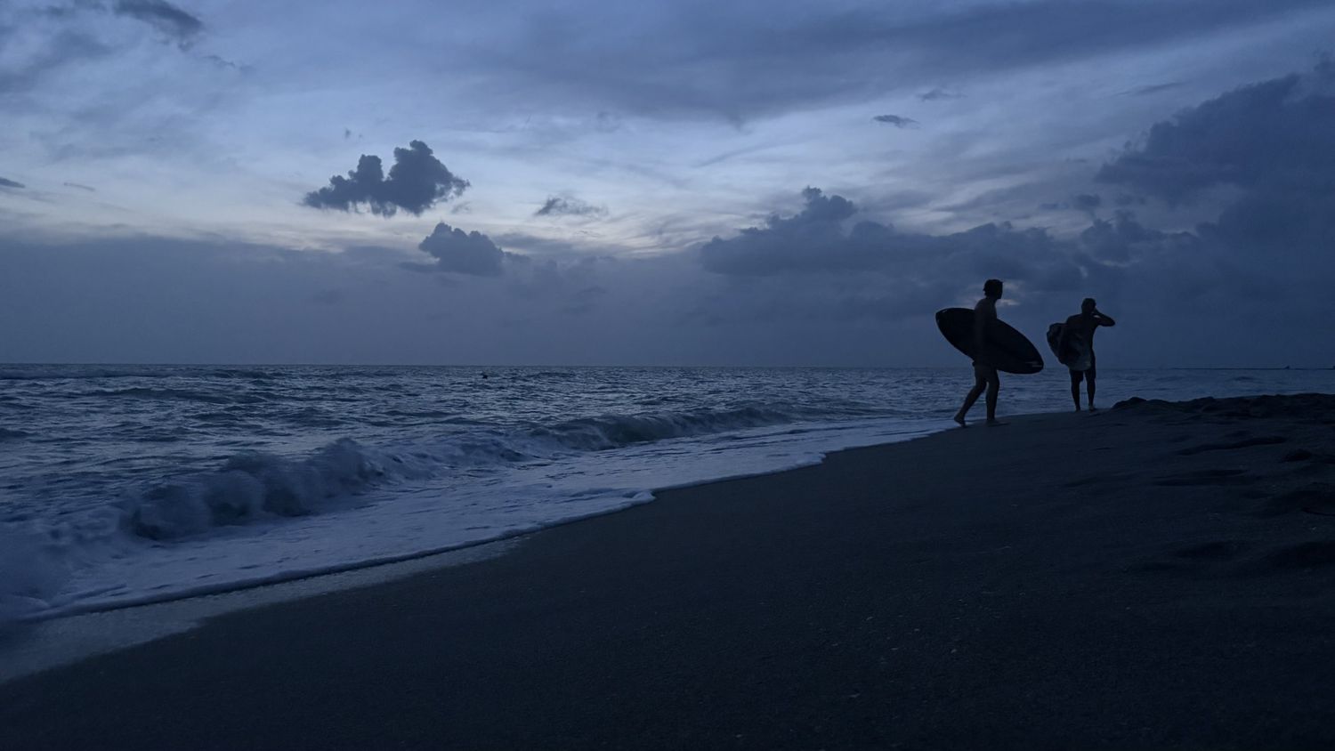 Two surfers carrying boards walk along a dark beach at dusk with dramatic cloudy skies and ocean waves.
