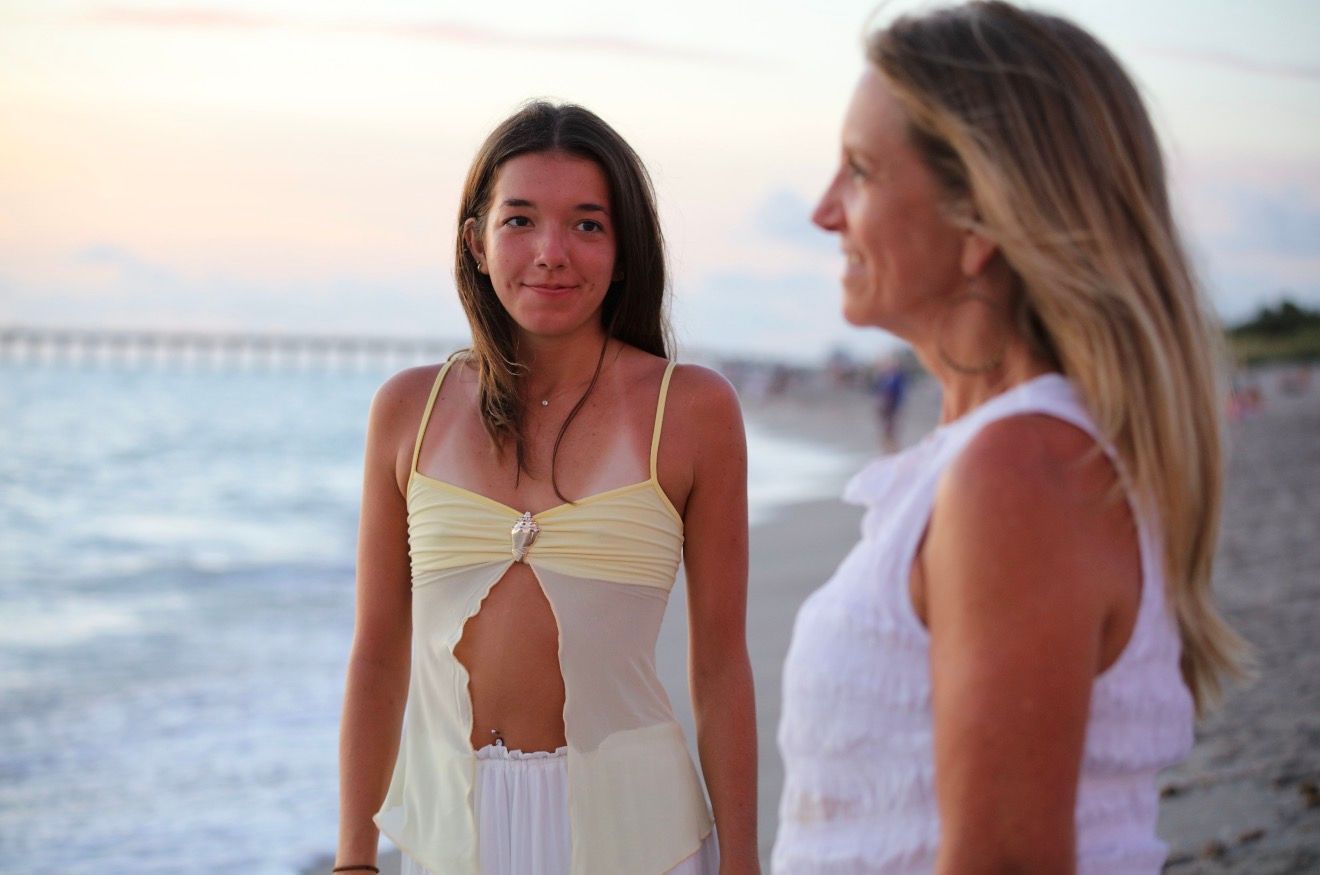 Two women in white dresses stand on a beach at sunset with a pier in the background.
