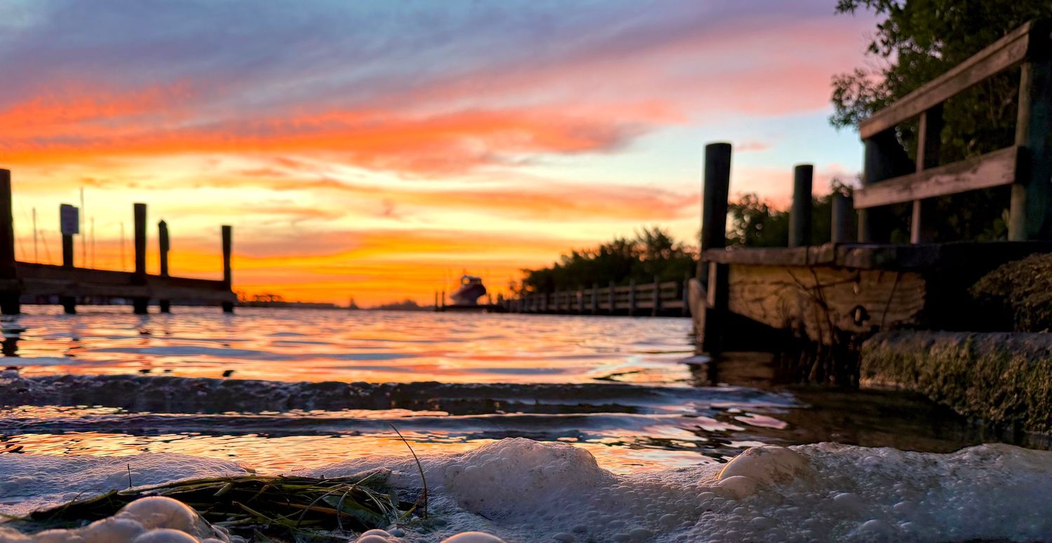 Stunning waterfront sunset with vibrant orange and purple skies reflecting on calm water near a weathered dock and pilings.