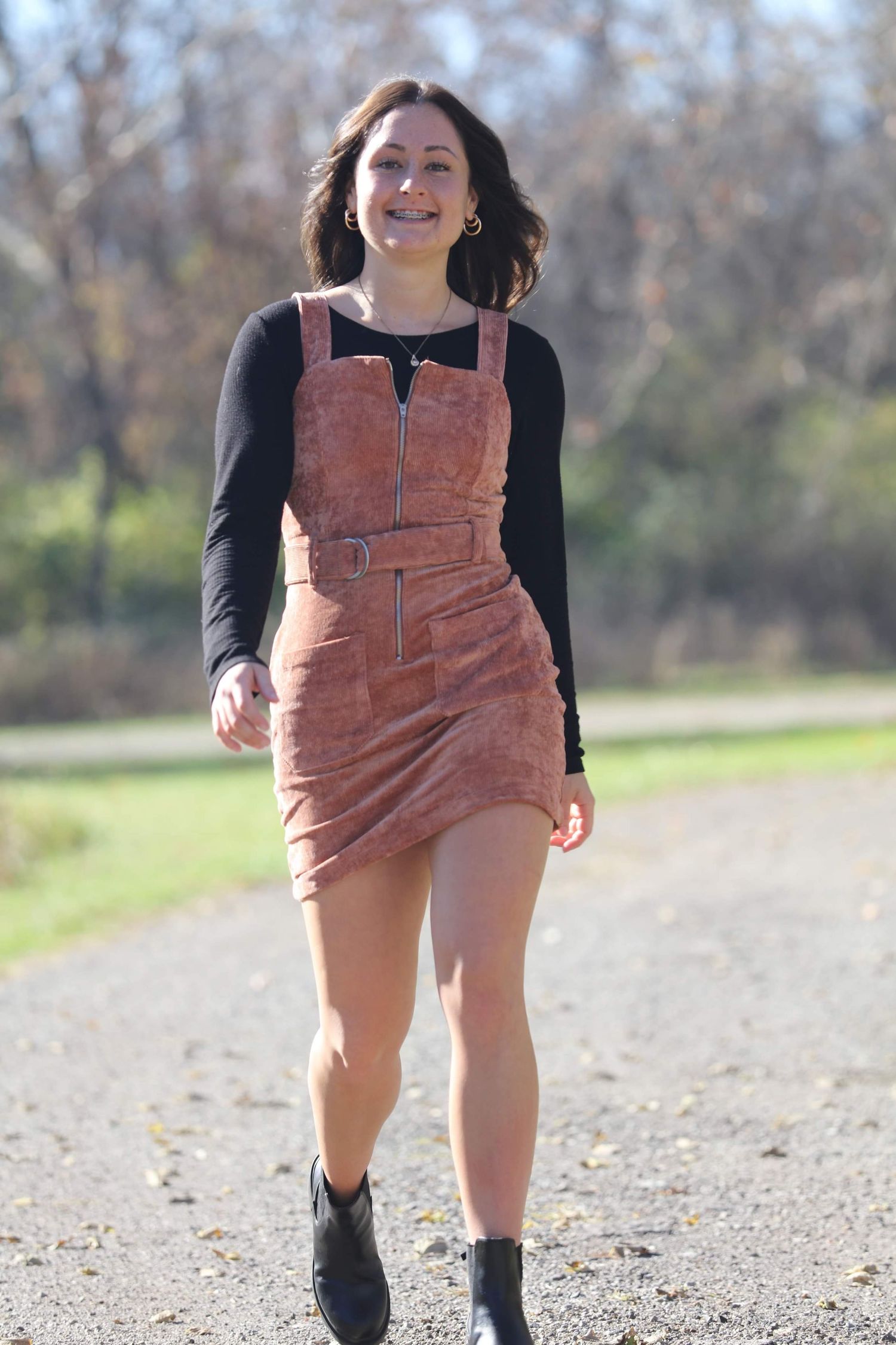 Smiling young woman walking on a gravel path wearing a pink corduroy overall dress with black long sleeve top and boots.
