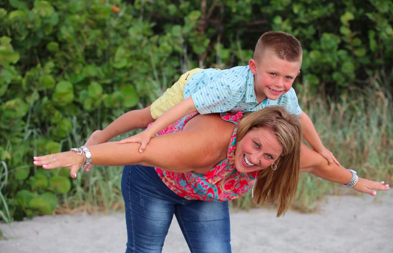 Woman smiling with arms outstretched while giving a young boy a piggyback ride on a sandy beach.