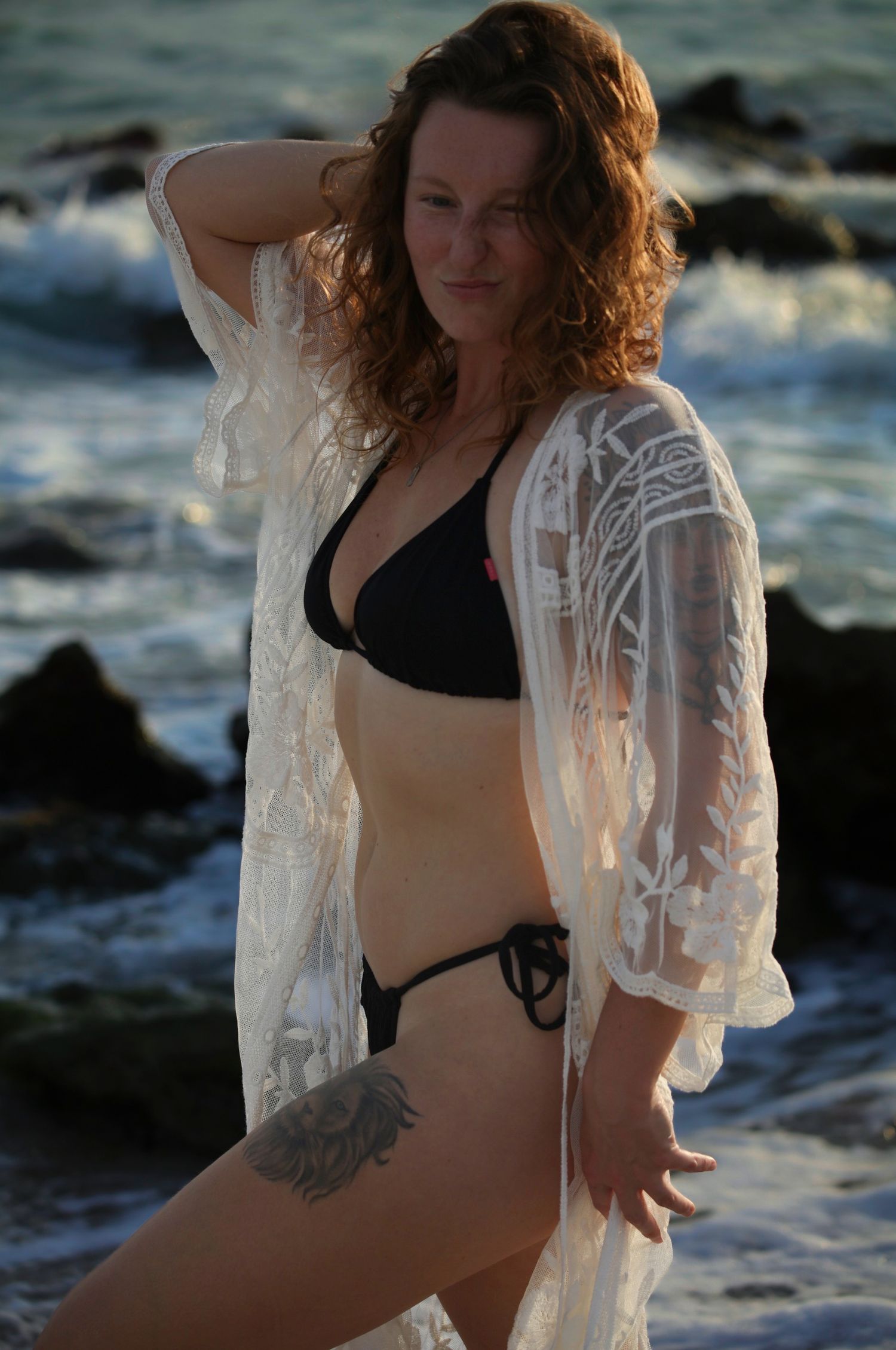 Woman with thigh tattoo in black bikini and white lace kimono poses on rocky beach at dusk.