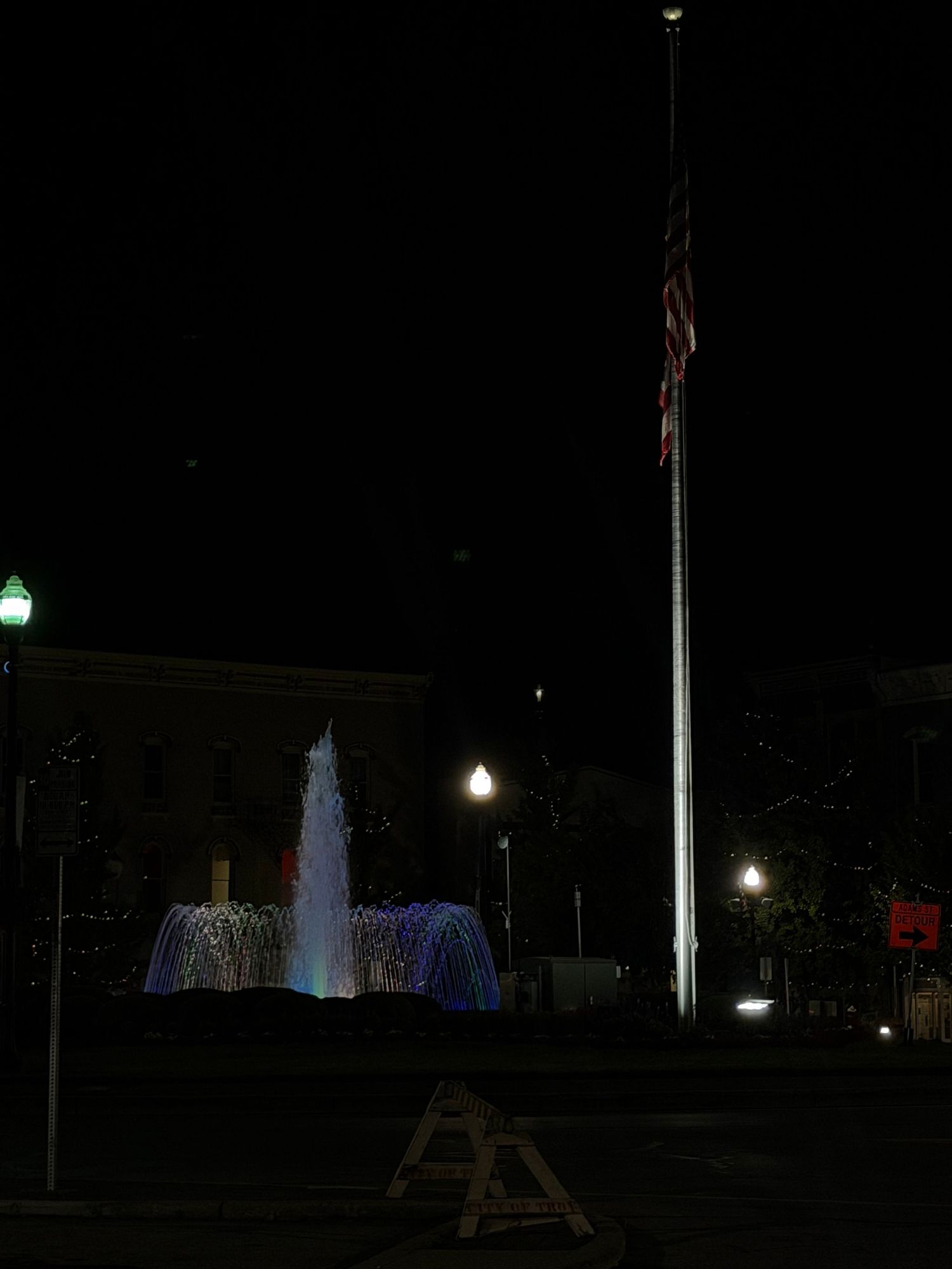 Illuminated fountain with colorful lights and American flag on tall pole at night in a public plaza.