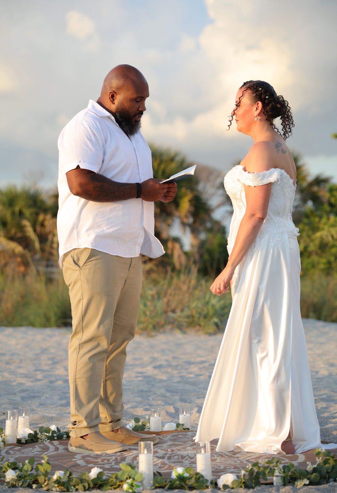 Couple exchanging vows on a sandy beach at sunset, groom in white shirt reading from paper, bride in elegant off-shoulder gown.