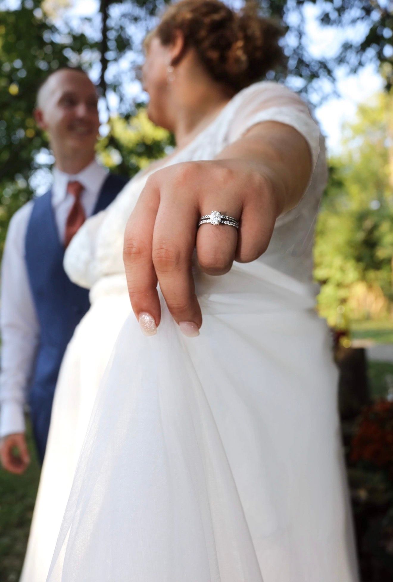 Bride in white gown shows off diamond engagement ring and wedding band while groom in blue vest smiles behind her outdoors.