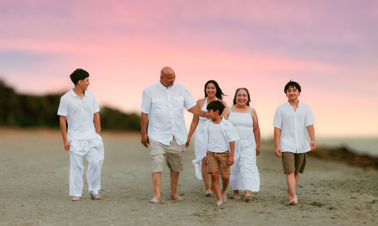 Family of six walks on beach at sunset with pink sky, wearing white and khaki outfits.