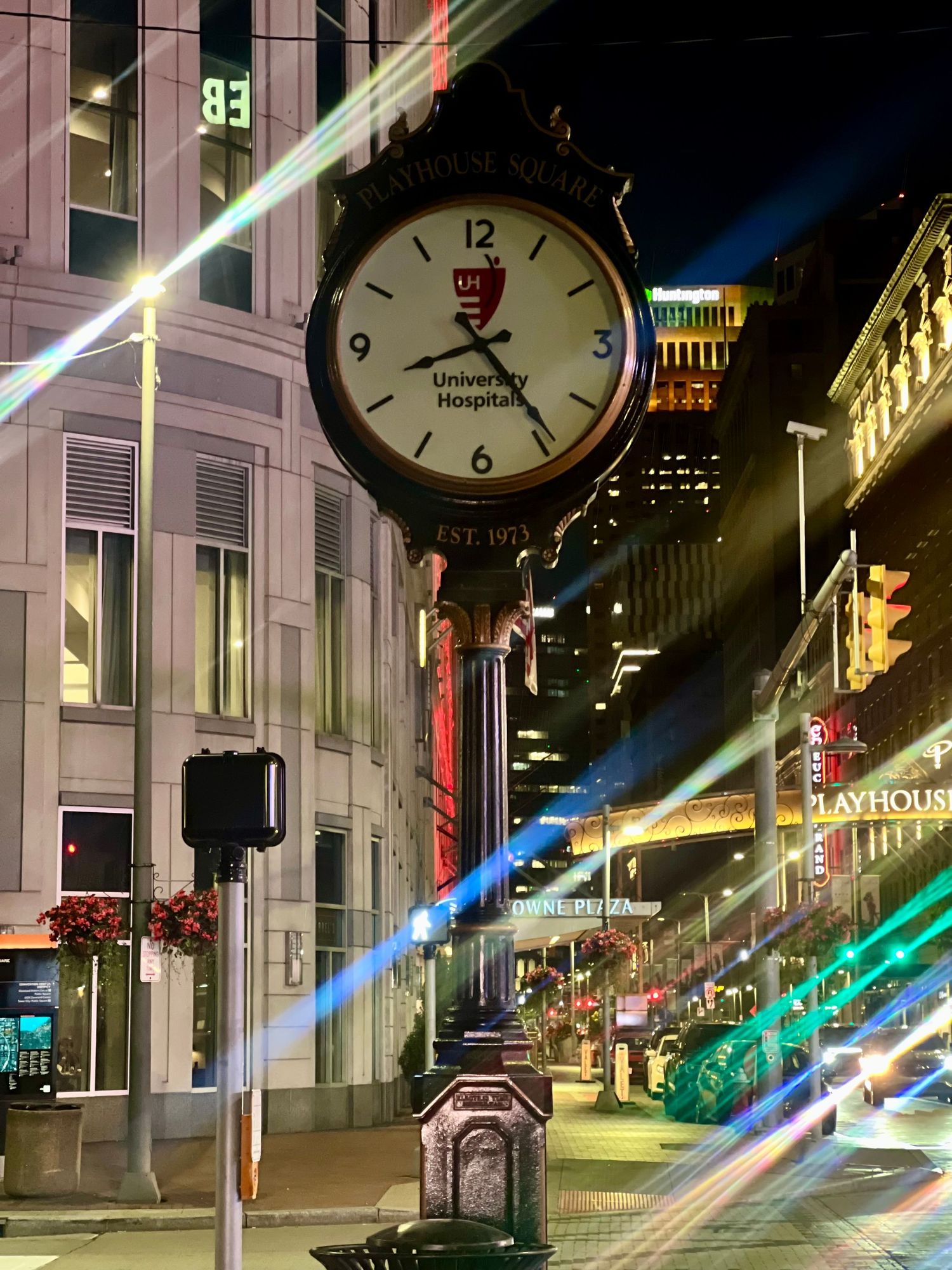 Night cityscape with a University Hospitals street clock glowing on a bustling urban street with light trails and illuminated buildings.