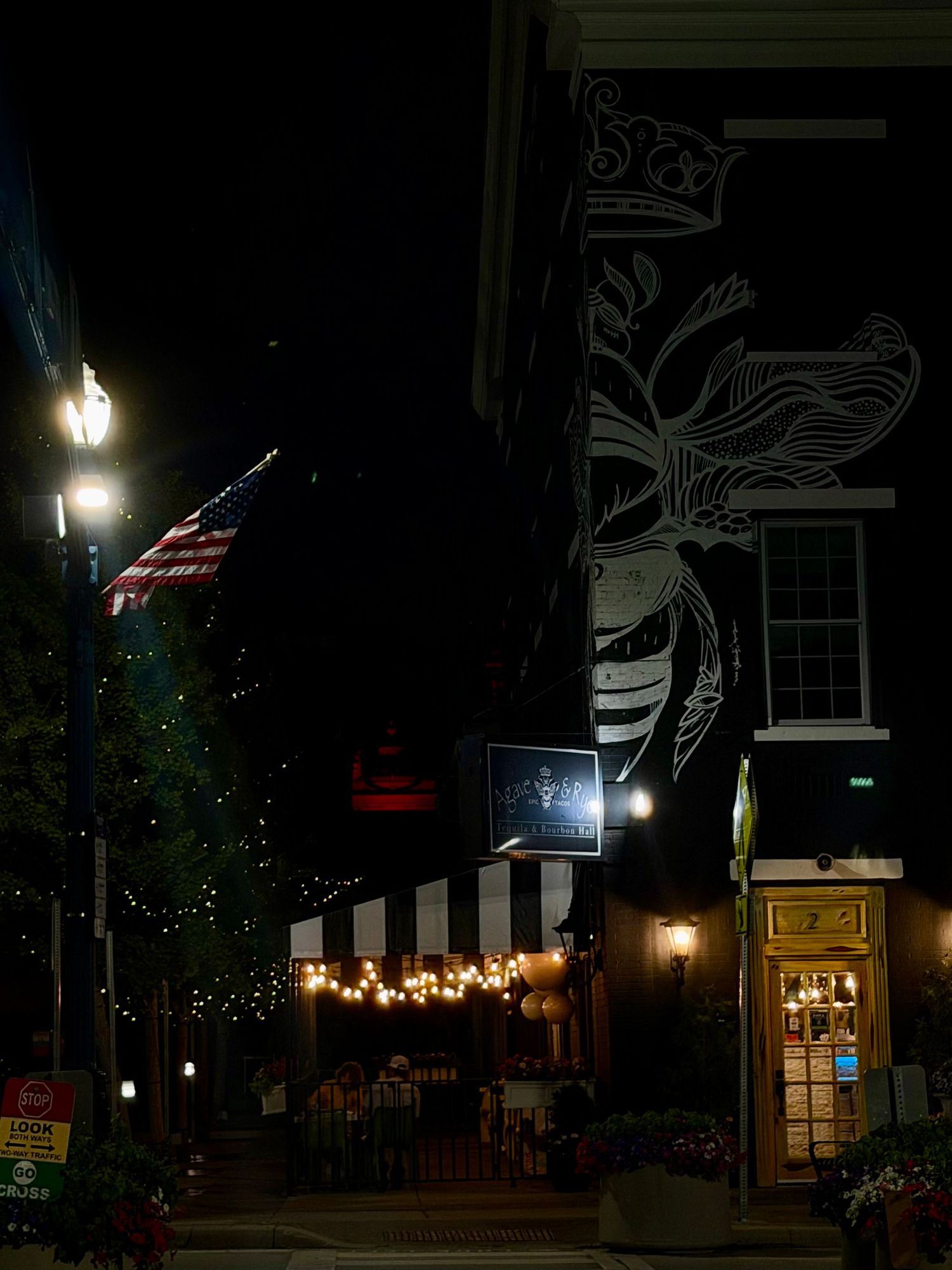 Nighttime street scene with American flag, string lights on striped awning, and decorative mural on brick building.