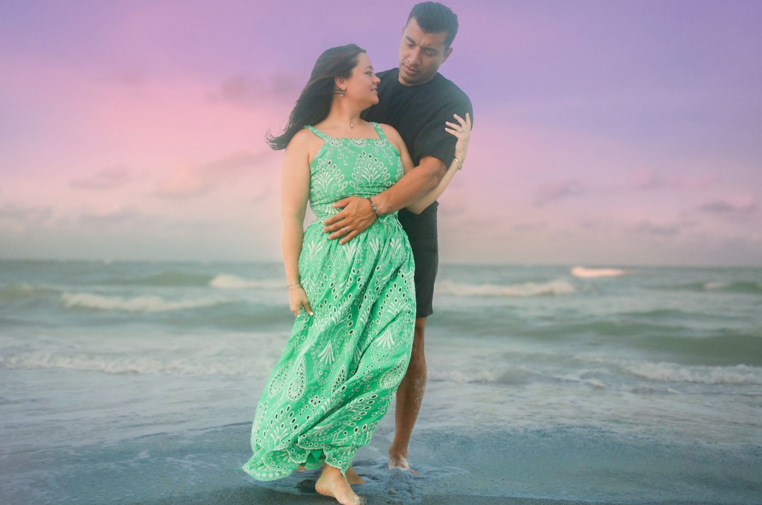 Couple embracing on a beach at sunset, woman in green maxi dress, waves and pink sky behind them.