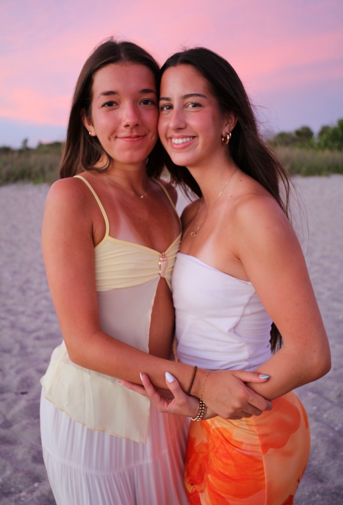 Two young women in summer dresses embrace on a beach at sunset with pink sky.