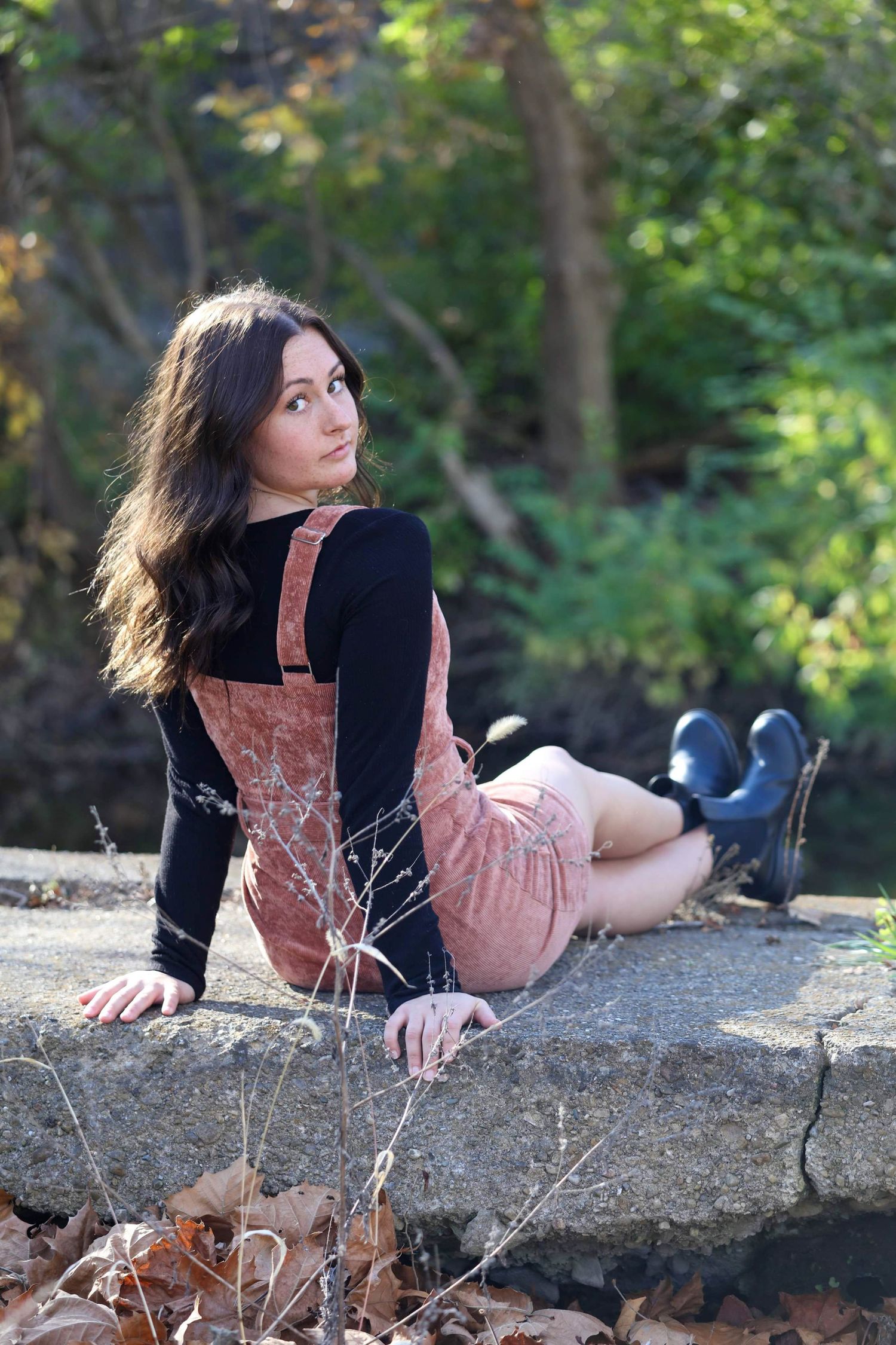 Young woman in pink overalls and black turtleneck sits on stone wall surrounded by autumn foliage in a park.