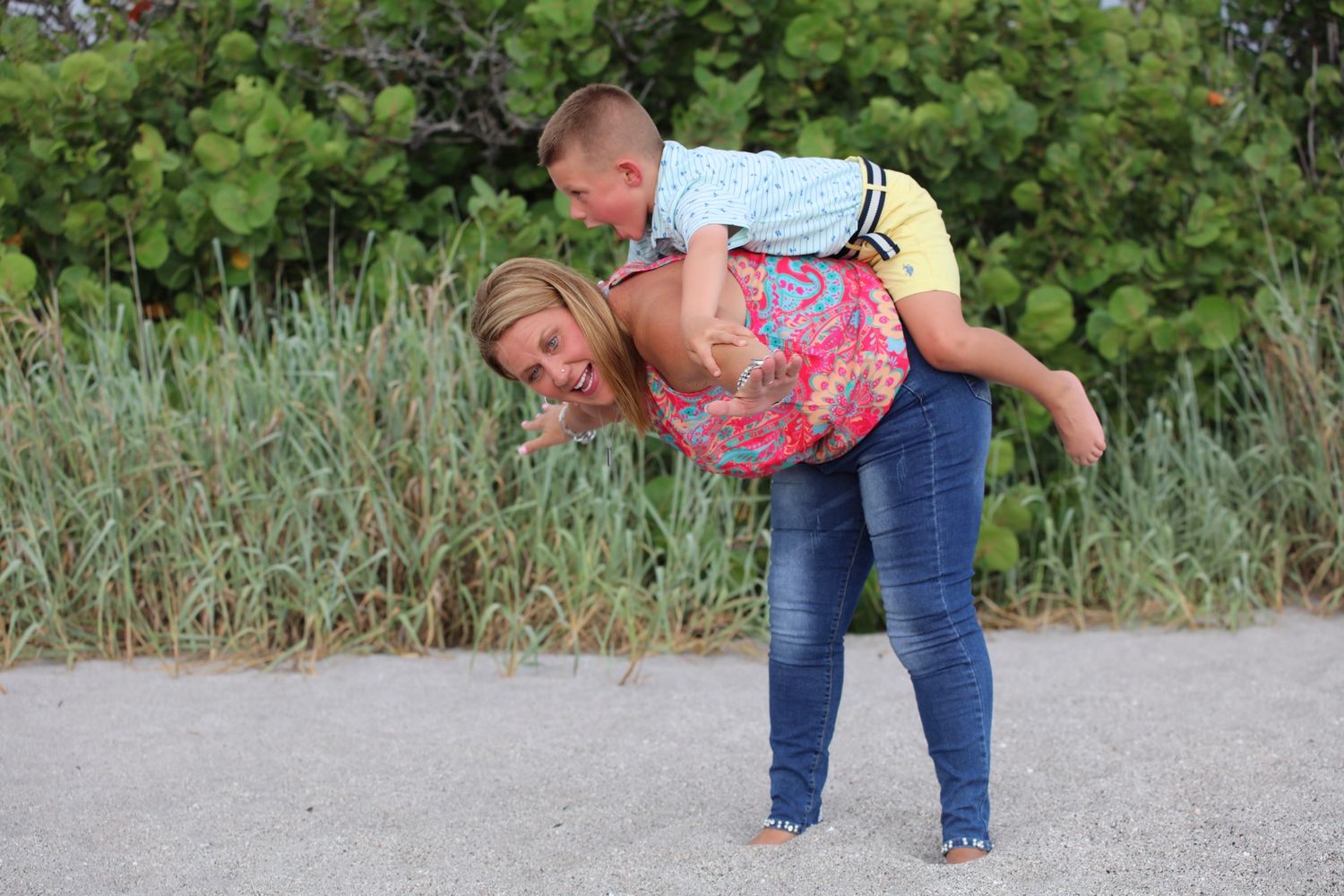 A woman gives a young boy a piggyback ride on a beach path with lush green vegetation in the background.
