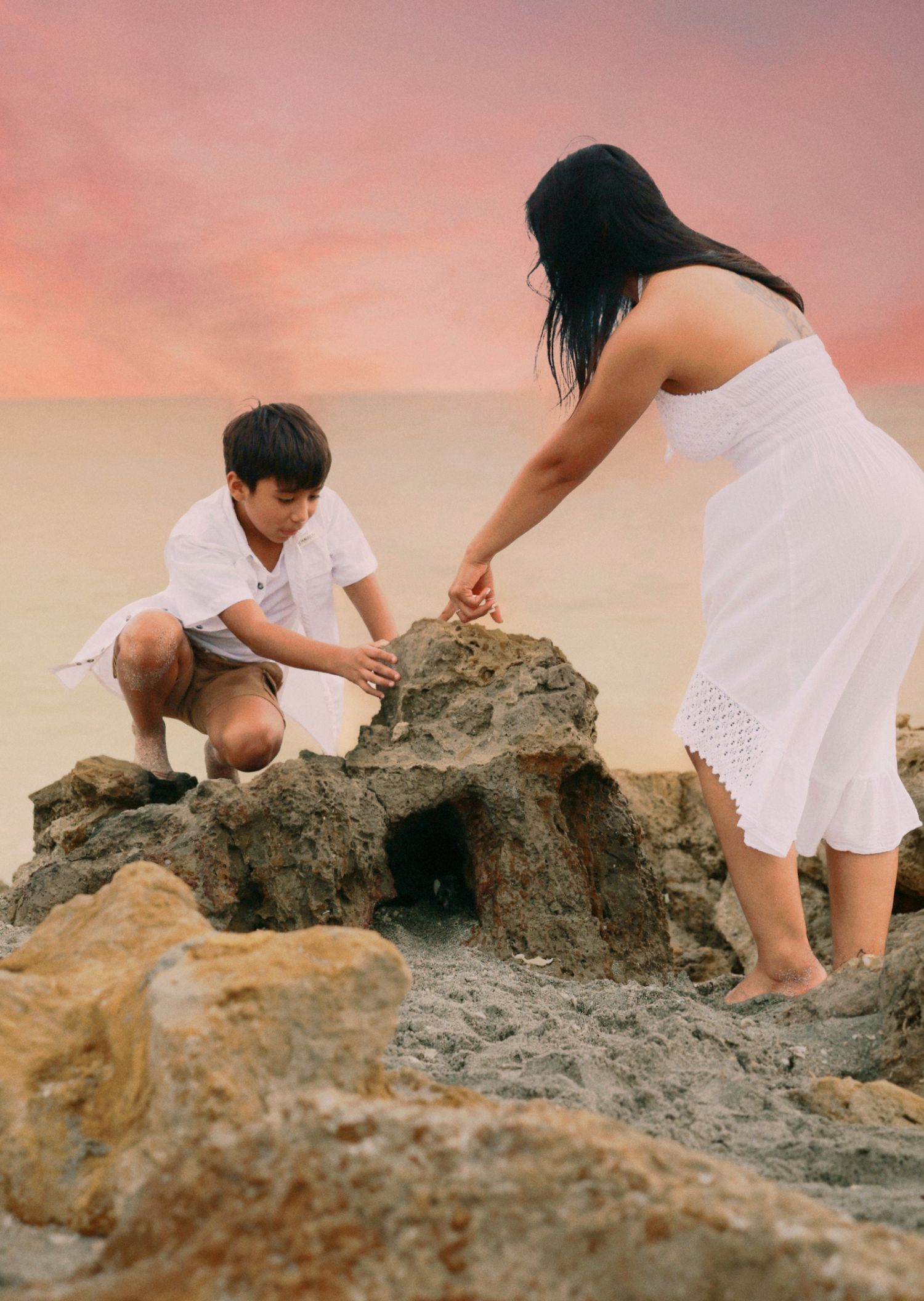 A woman in white dress and young boy explore rocky formations together on a beach at sunset.