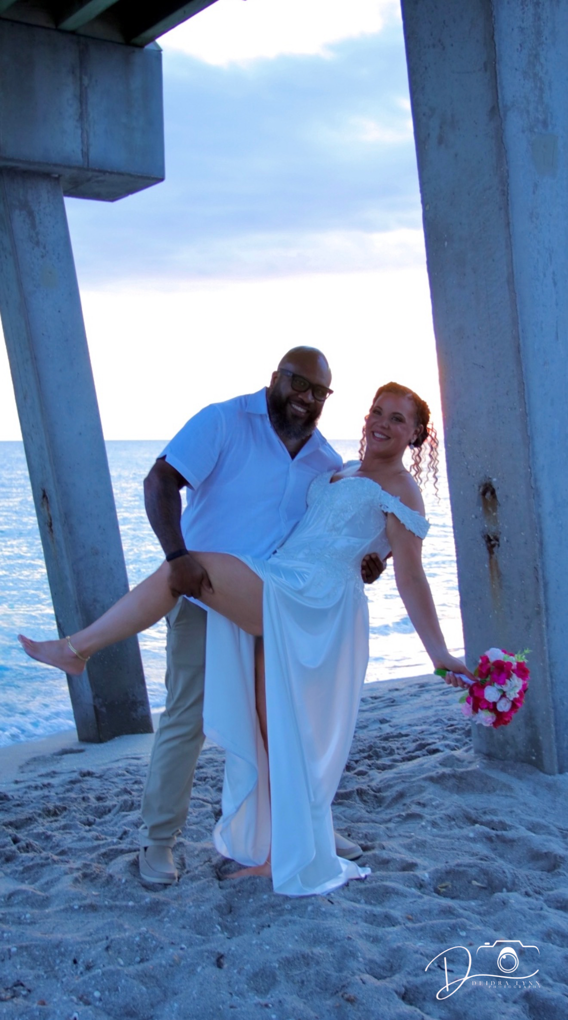 A joyful interracial couple poses at beach wedding under pier, bride in white gown holding pink bouquet, groom lifting her leg.