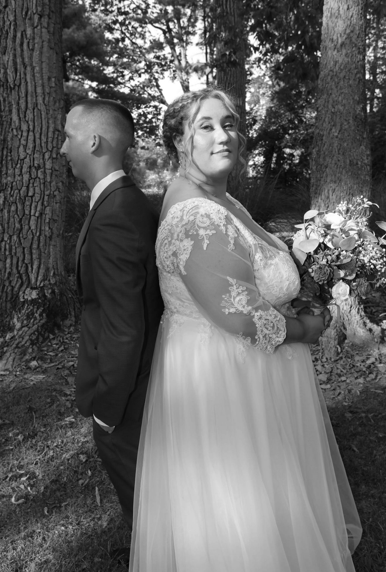Black and white wedding photo of bride in lace gown holding bouquet standing back-to-back with groom among trees.