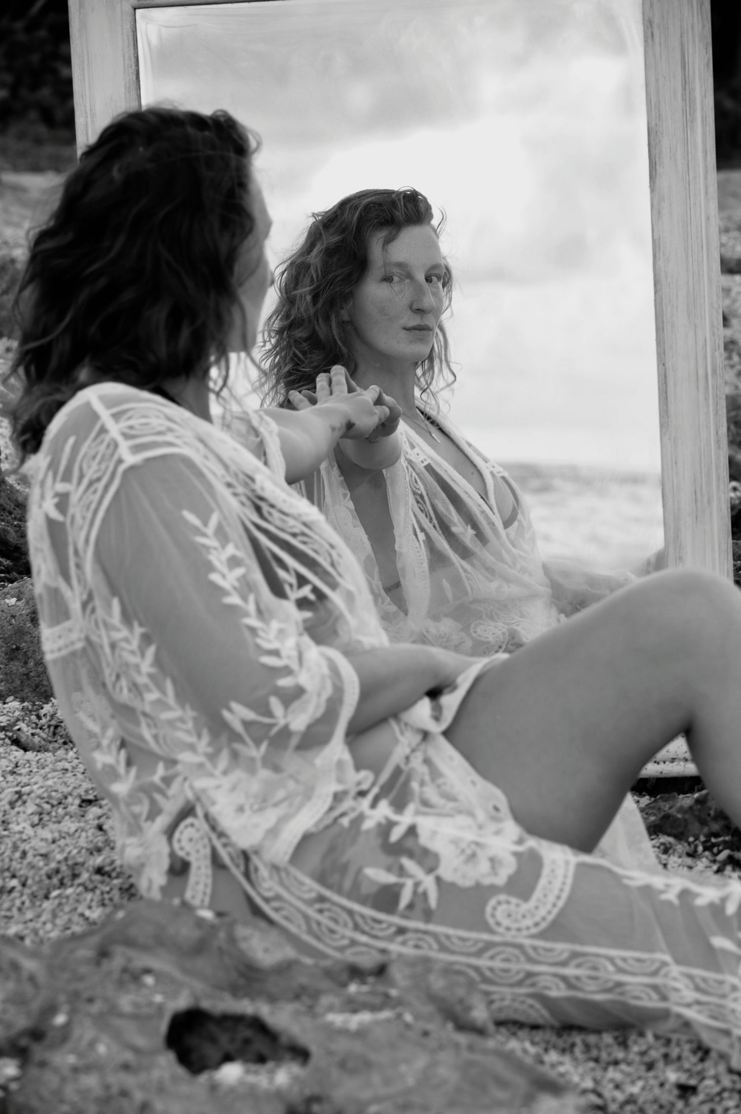 Black and white photo of a woman in lace robe sitting outdoors before a mirror reflecting her image near the ocean.
