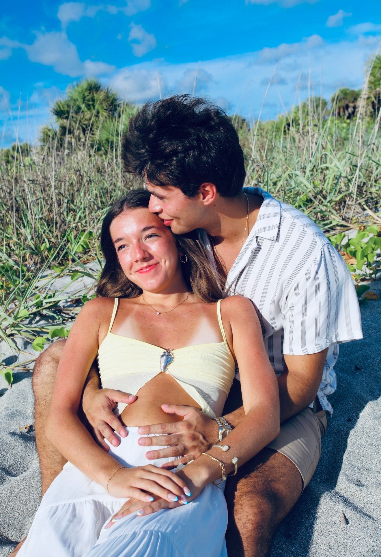 Couple sitting on beach sand, woman in yellow bikini top and white skirt, man kissing her head during maternity shoot.