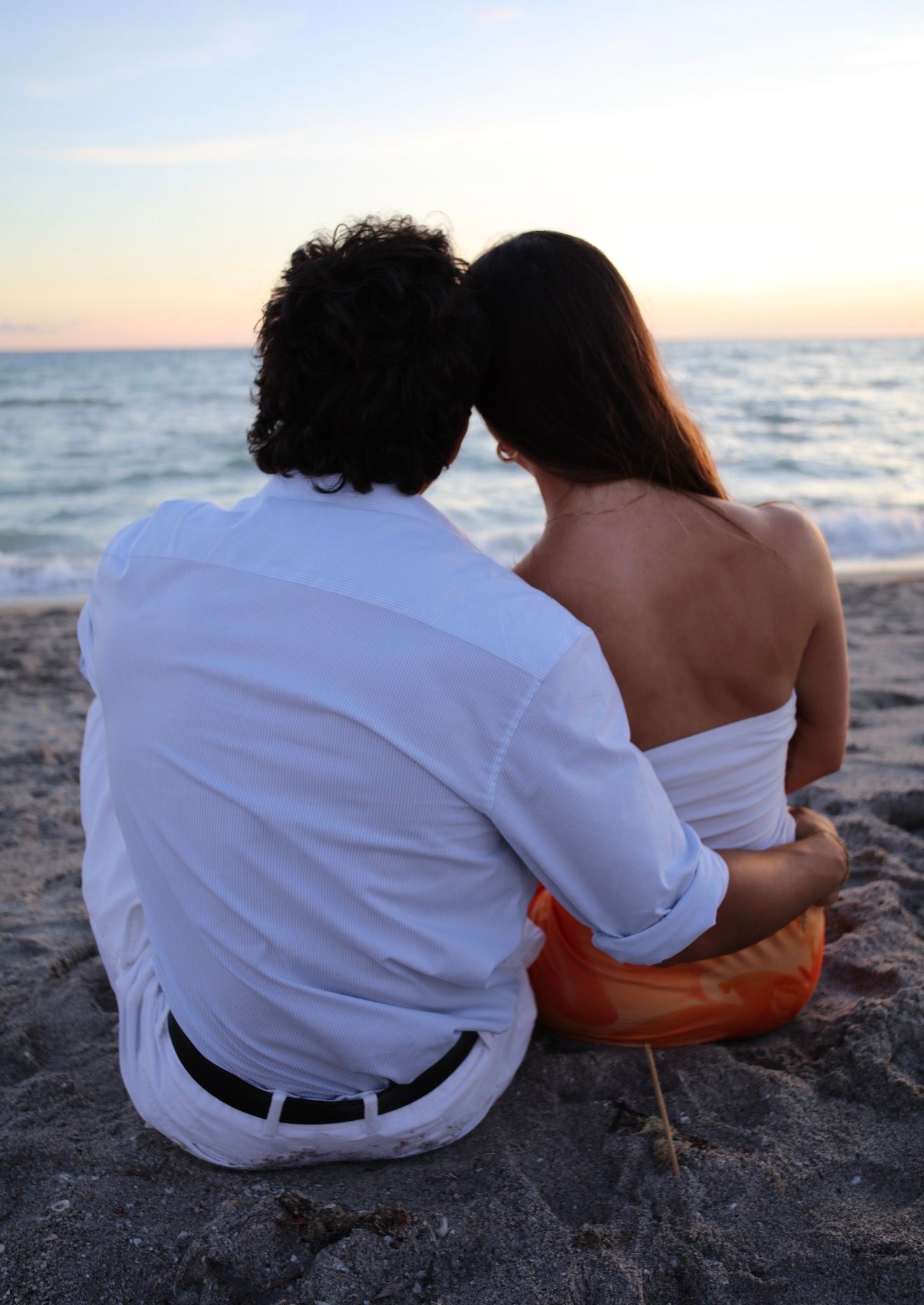 A couple sits closely together on a sandy beach, leaning heads together while watching the sunset.