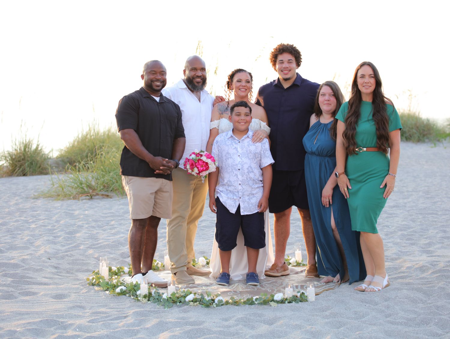 Group of seven people posing together on a sandy beach, smiling for a photo during a casual outdoor gathering.