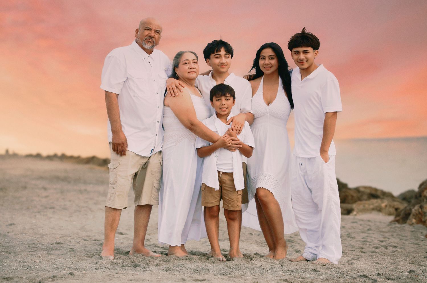 Family of six posing together on a beach at sunset, all wearing white outfits, with a vibrant pink sky backdrop.