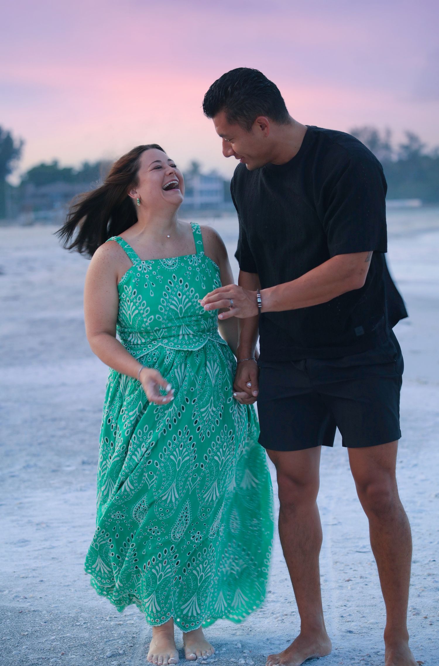 Couple laughing together on beach at sunset, woman in green dress.
