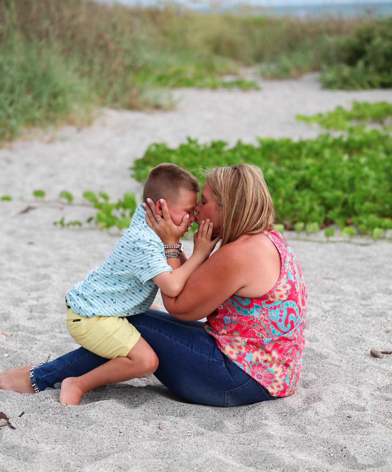 A woman kisses a young boy on a sandy beach, both sitting on the ground near coastal vegetation.