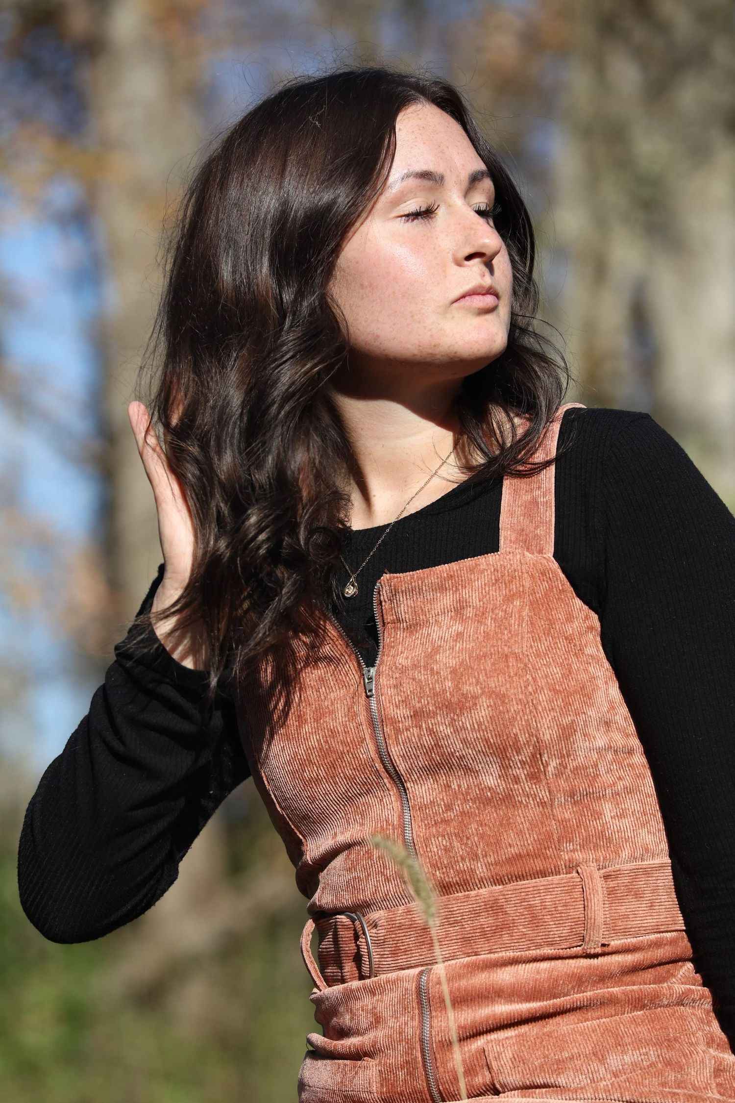 Young woman with dark hair wearing a rust corduroy overall dress over black top, posing outdoors.
