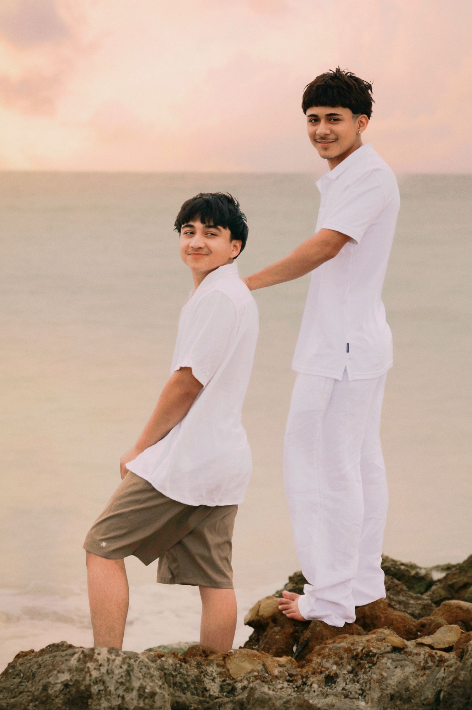 Two teenage boys in white shirts and khaki shorts posing on rocks at a pastel sunset beach.