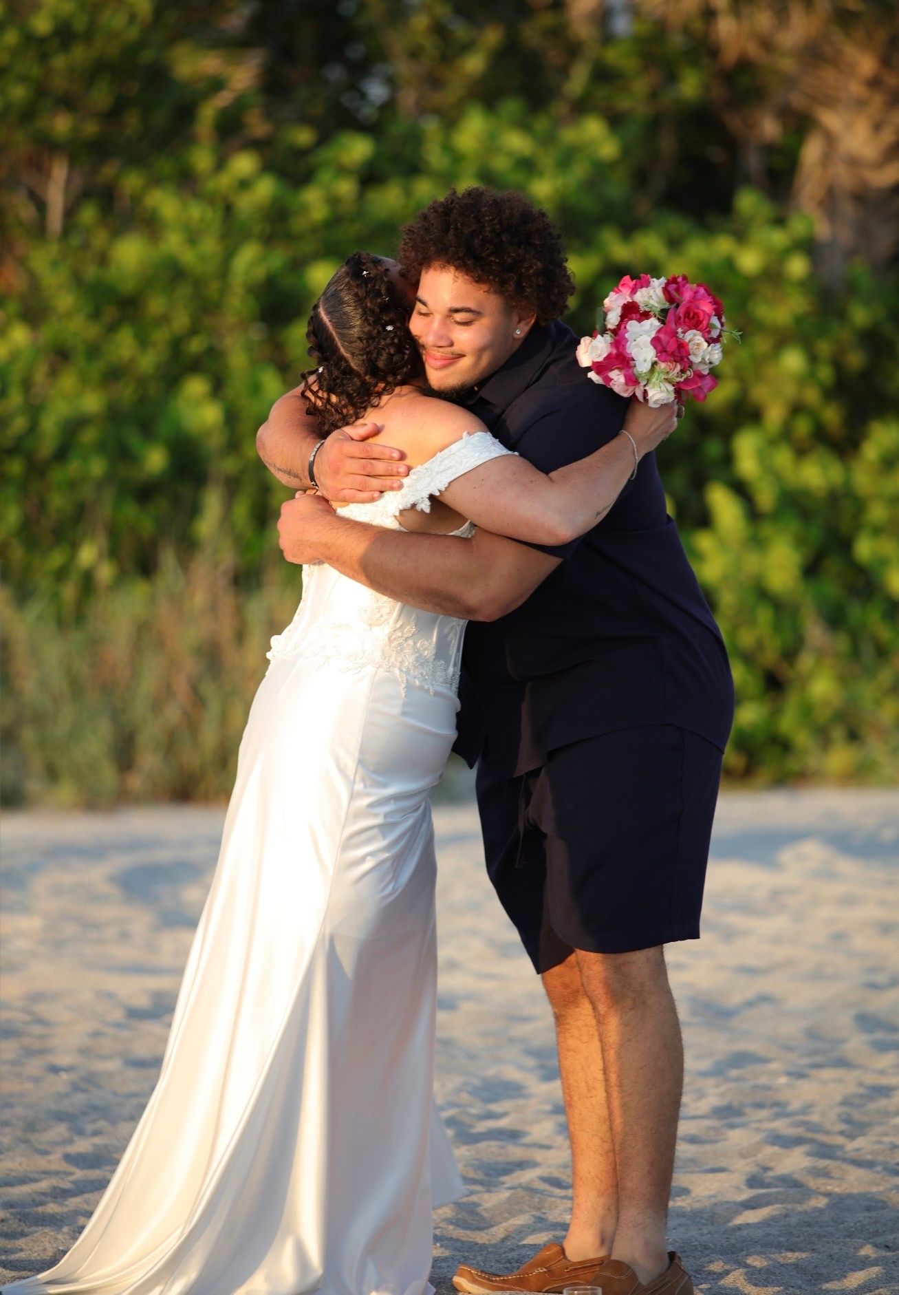 Bride in white gown embraces guest in navy outfit on sandy beach during outdoor wedding ceremony.