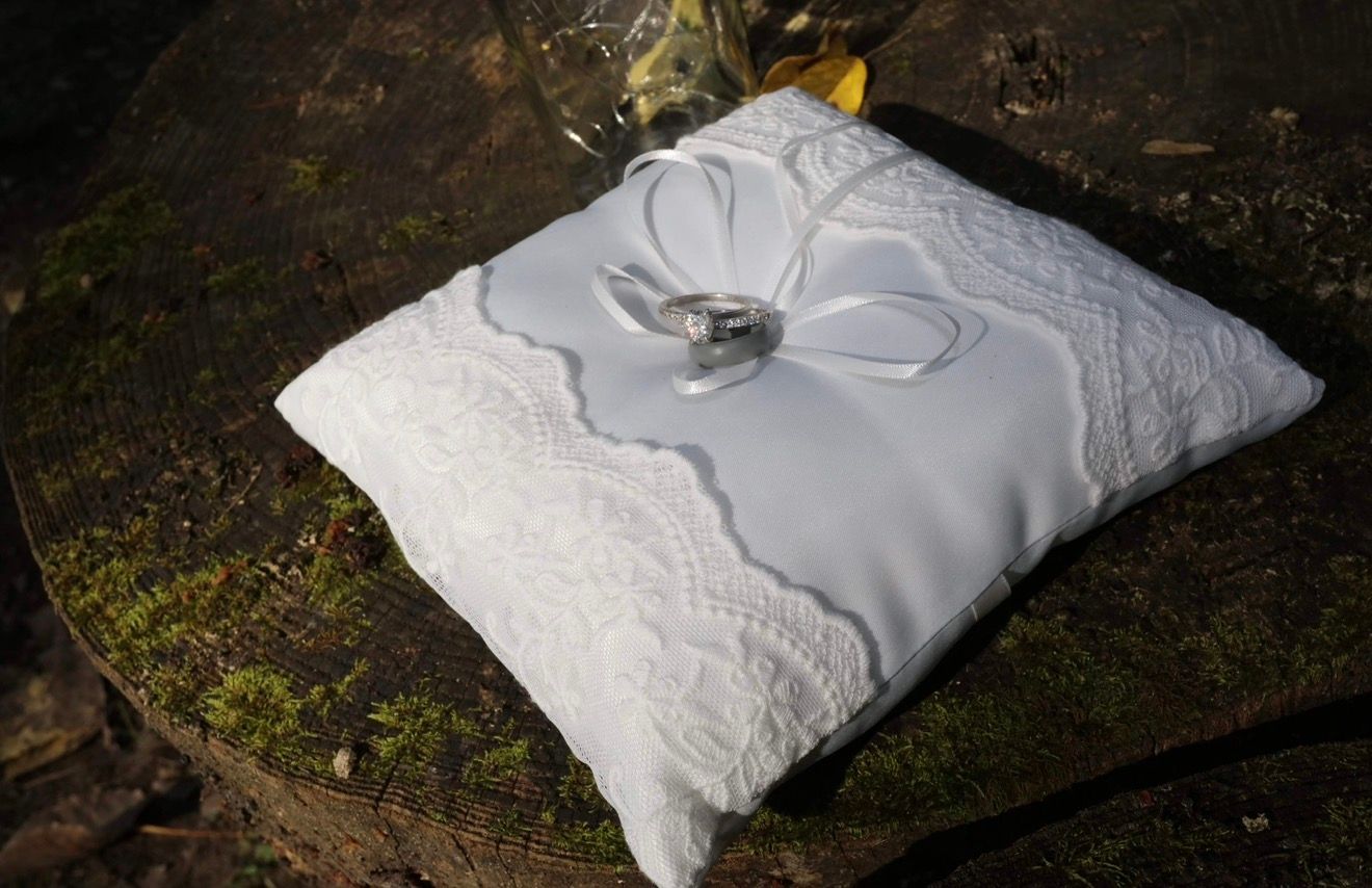 White satin ring bearer pillow with lace trim and diamond wedding rings tied with a bow, displayed outdoors on moss.