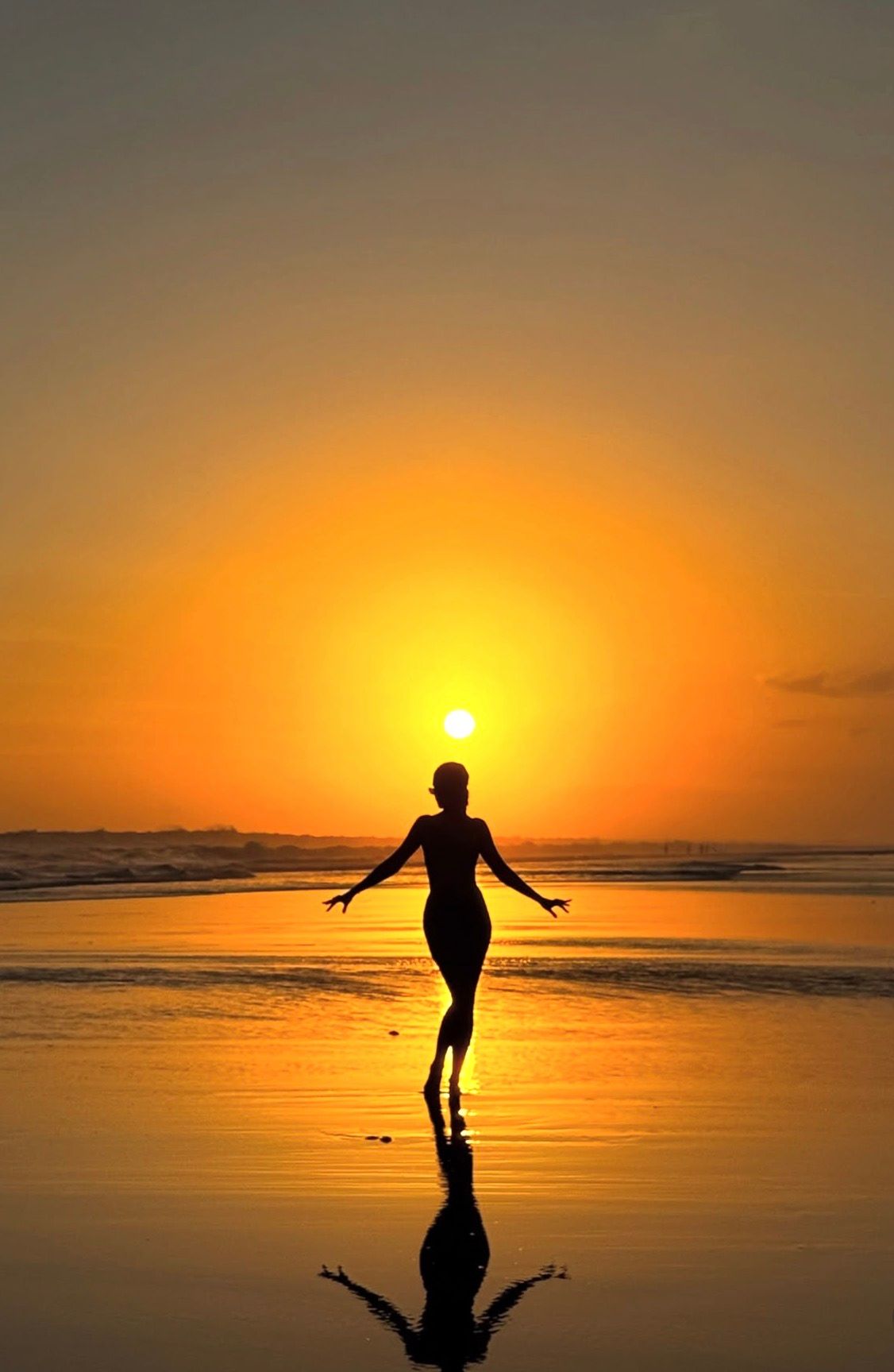 Silhouette of a woman dancing on a beach at sunset with golden orange sky and sun reflected on wet sand.