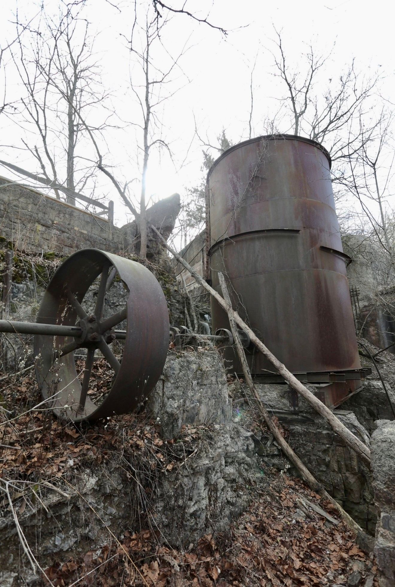 Rusty abandoned industrial equipment with a large wheel and metal tank on a rocky hillside surrounded by bare trees.