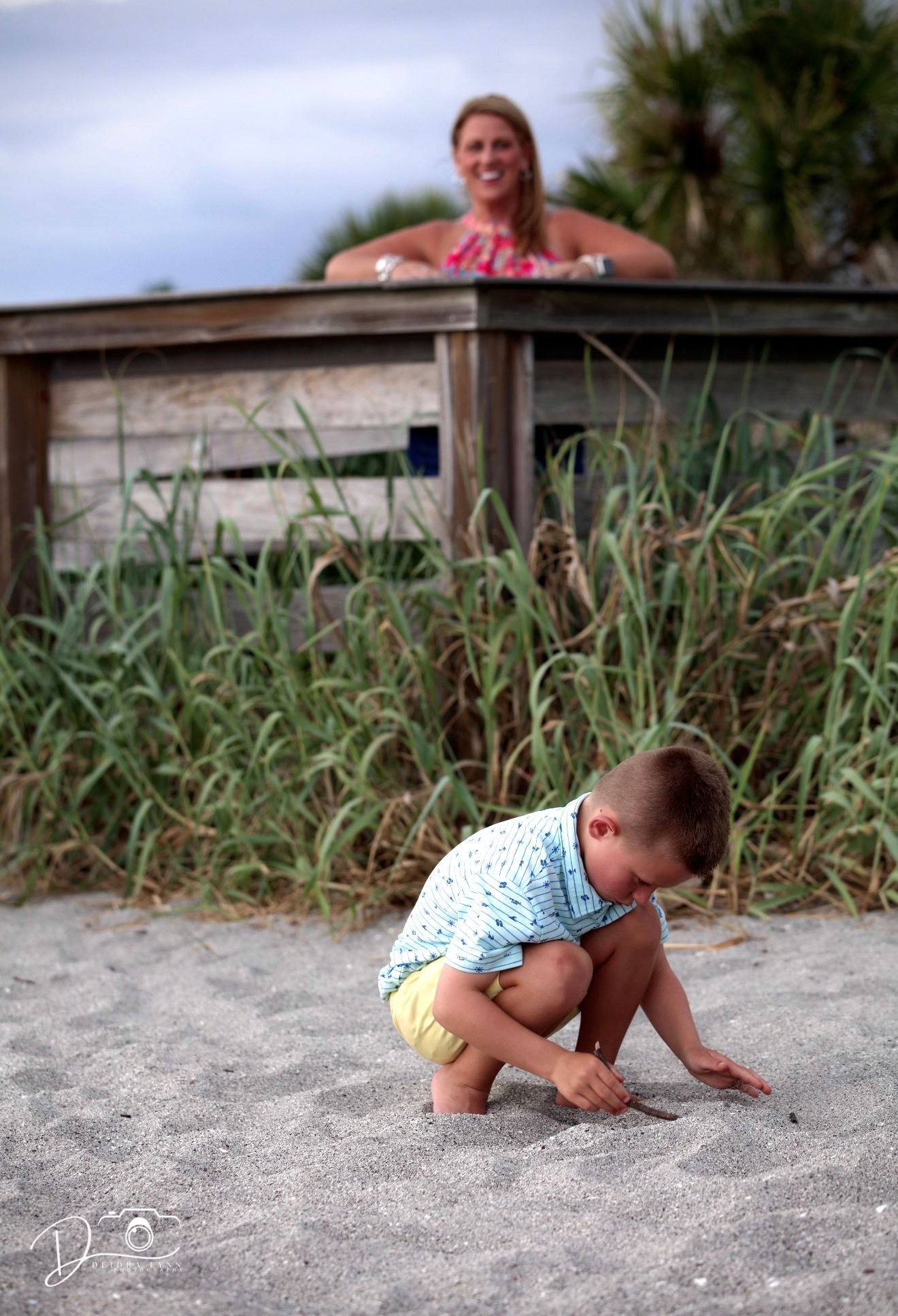 A toddler plays in beach sand while a smiling woman watches from a wooden deck surrounded by tall green grass.