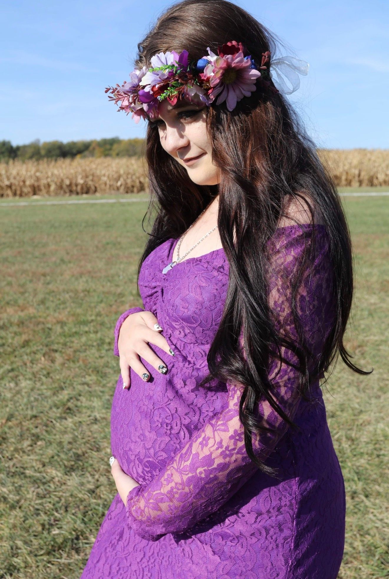 Pregnant woman in purple lace off-shoulder dress with floral crown poses outdoors in green field near cornfield.