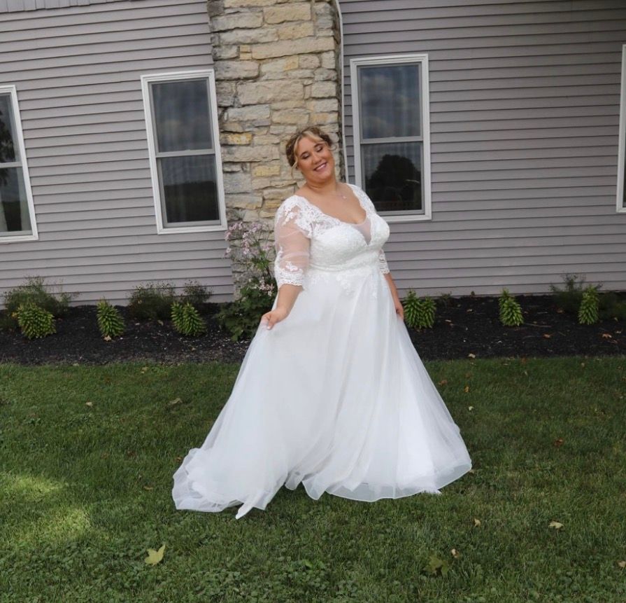 Woman in white lace wedding gown with tulle skirt posing outdoors on green lawn beside stone building.