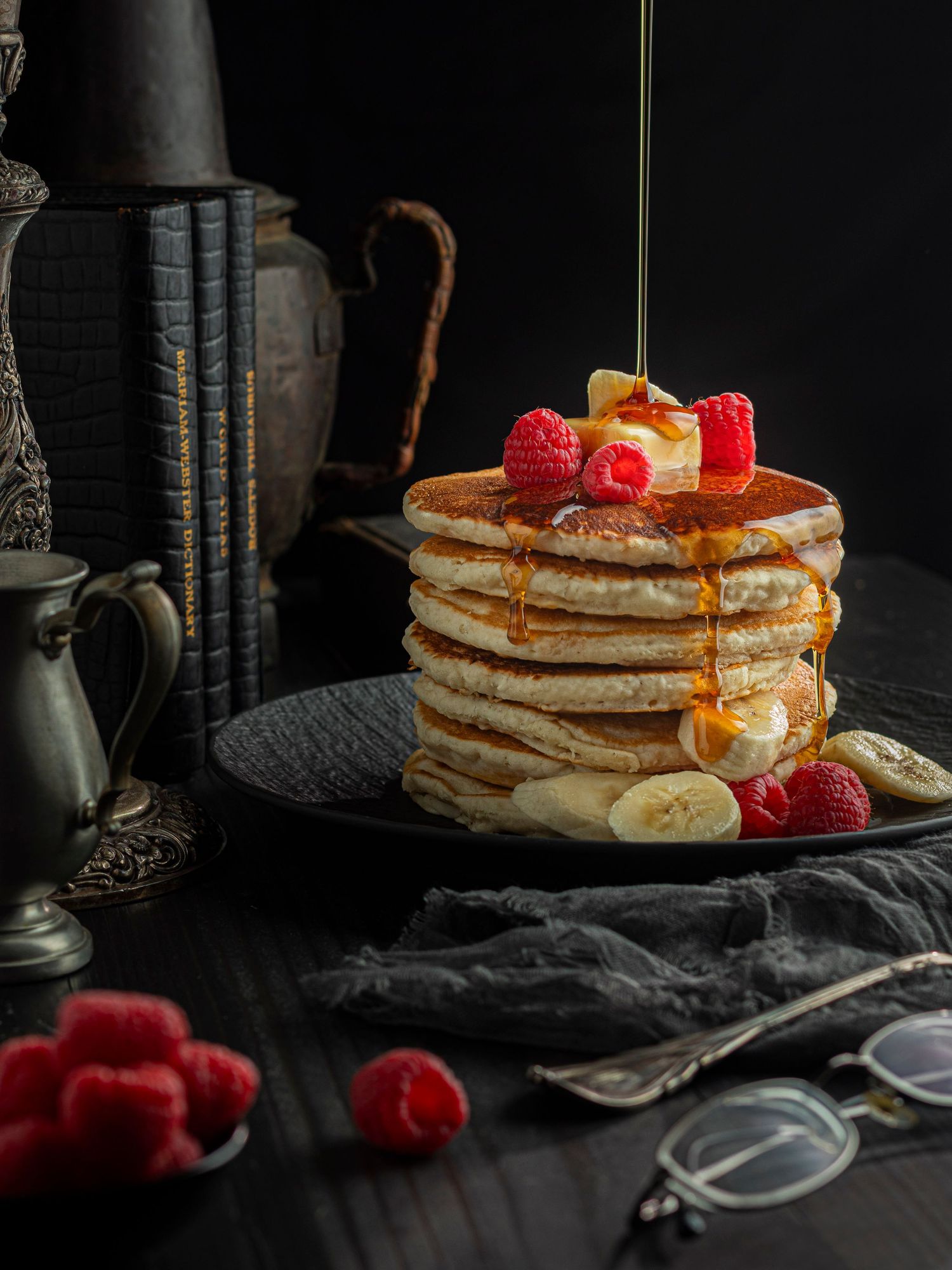 Head-on shot of a stack of buttermilk pancakes with bananas, berries and a drizzle of maple syrup being poured over the top