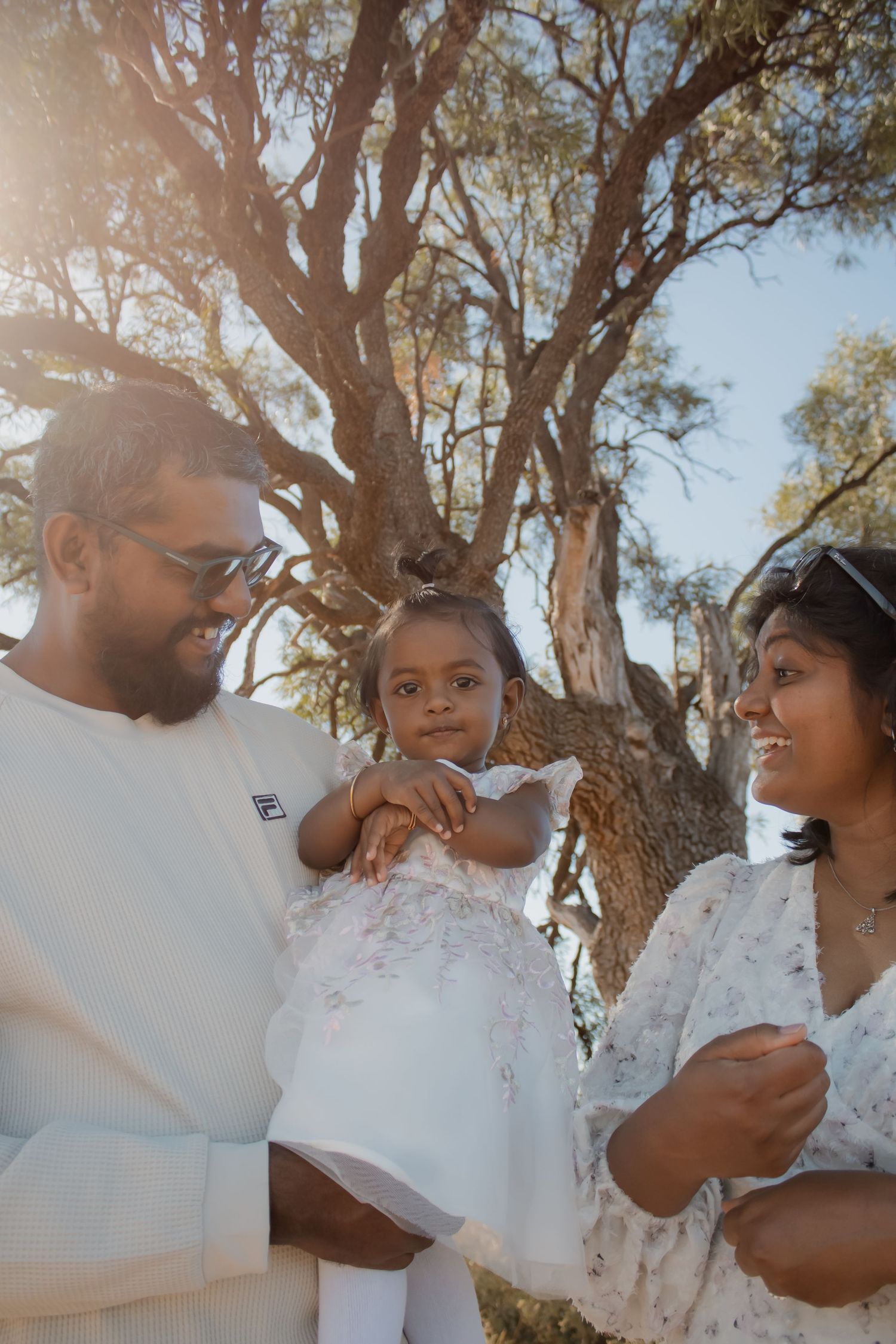 A joyful family of three outdoors, parents holding their young daughter in white dress, bathed in golden sunlight near trees.