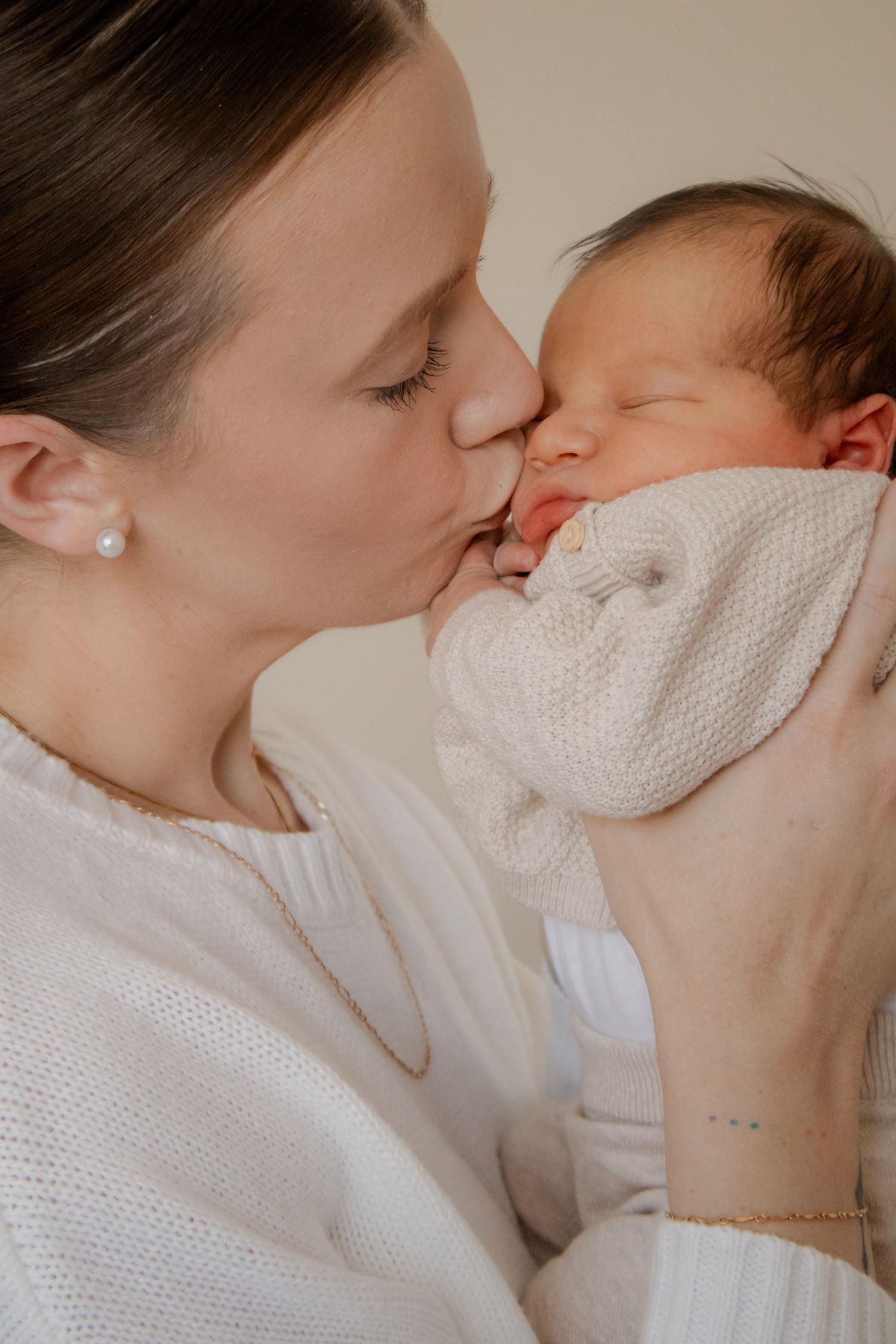 A mother tenderly kisses her sleeping newborn baby wrapped in a white knit blanket.