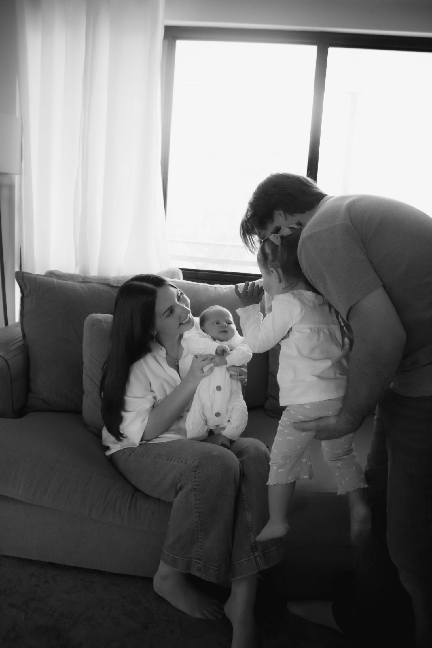 Black and white family photo of mother holding newborn on sofa while father lifts toddler nearby.