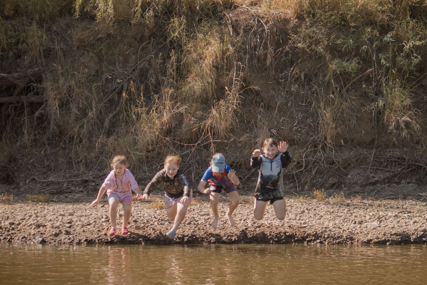 Four children playing and splashing along a muddy riverbank on a sunny day.