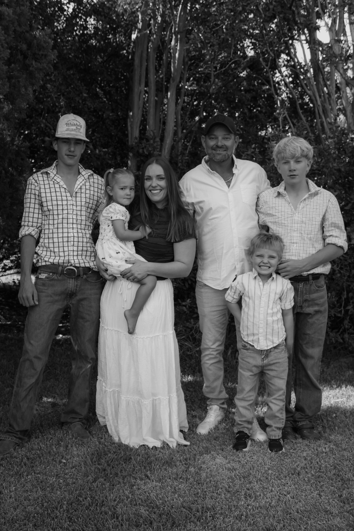 Black and white family portrait of six people posing outdoors on grass with trees in the background.