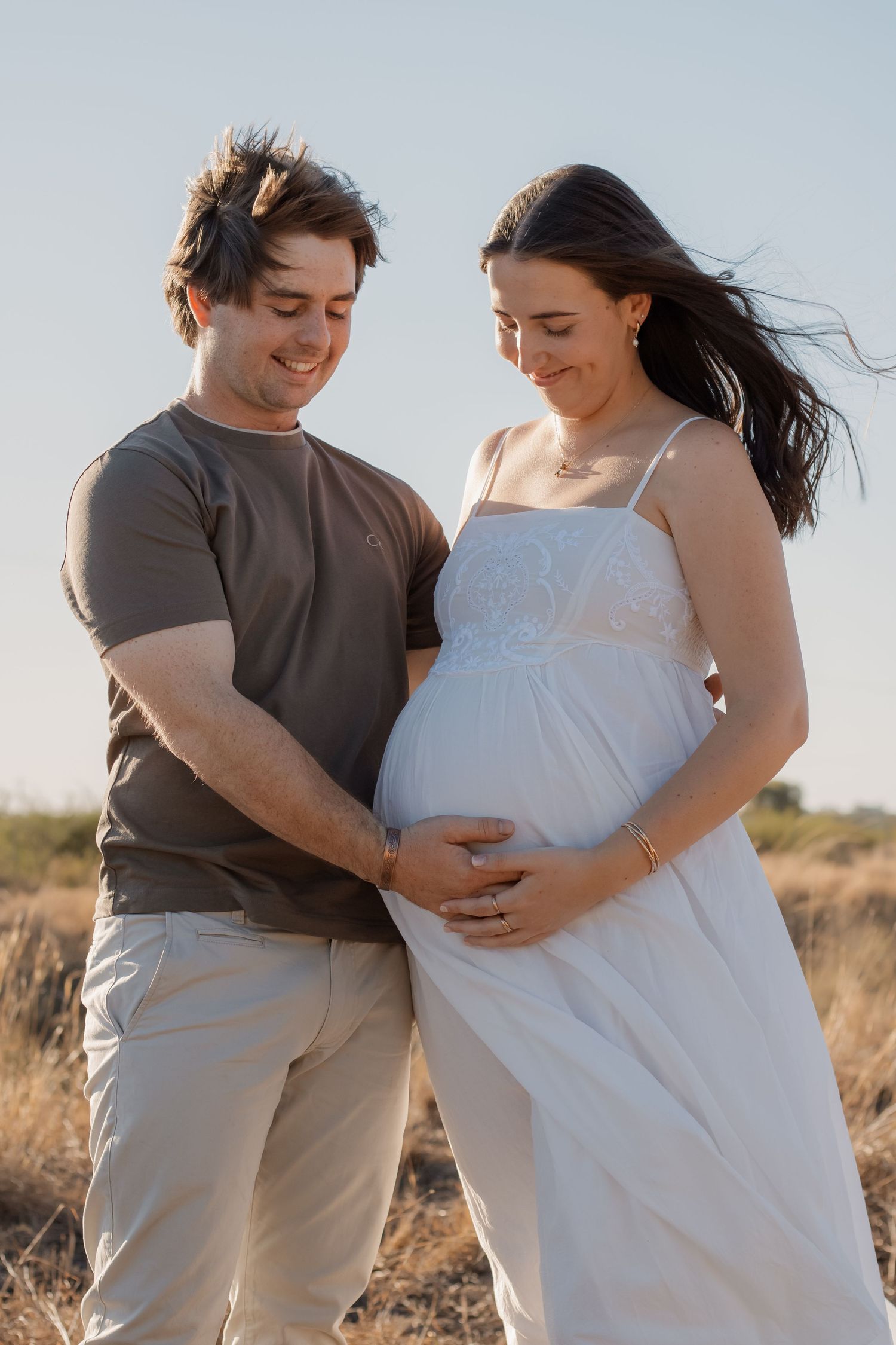 A couple smiling and cradling a pregnant belly outdoors in a sunny field, the woman in a white dress.