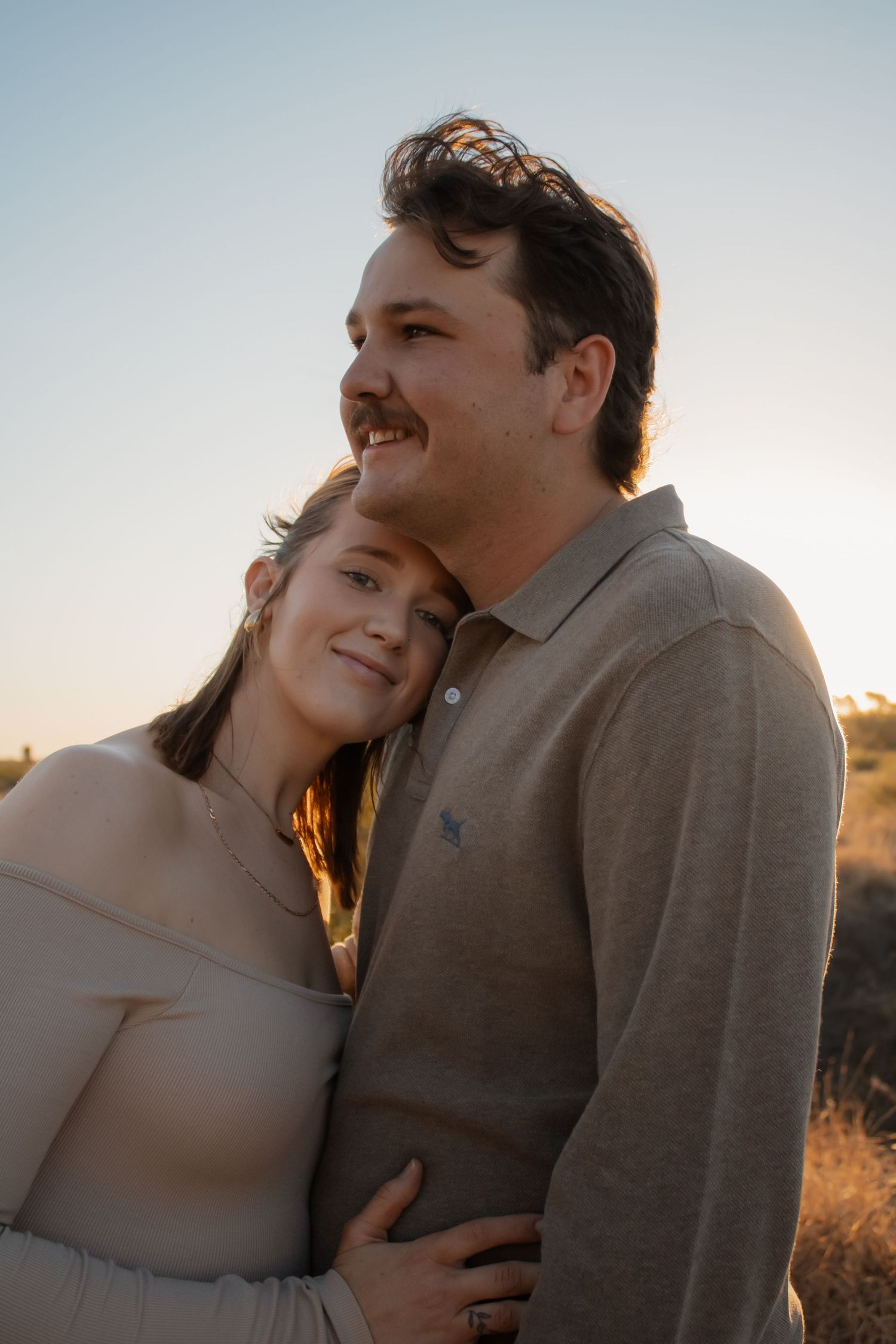 A couple embraces at golden hour, woman resting head on man's shoulder, smiling outdoors in a field.