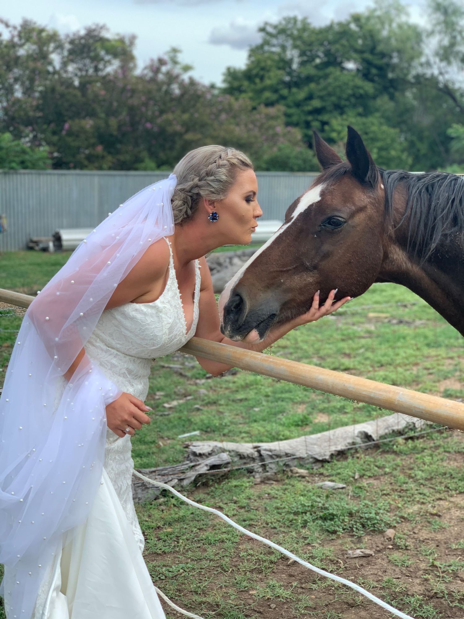 Bride in white wedding gown and veil sharing a tender moment with a horse at a farm.