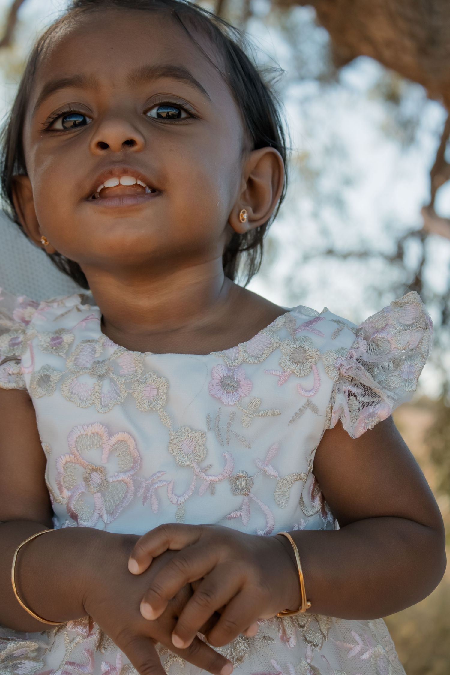 Adorable toddler girl in floral dress with gold bracelet and earrings smiling outdoors.