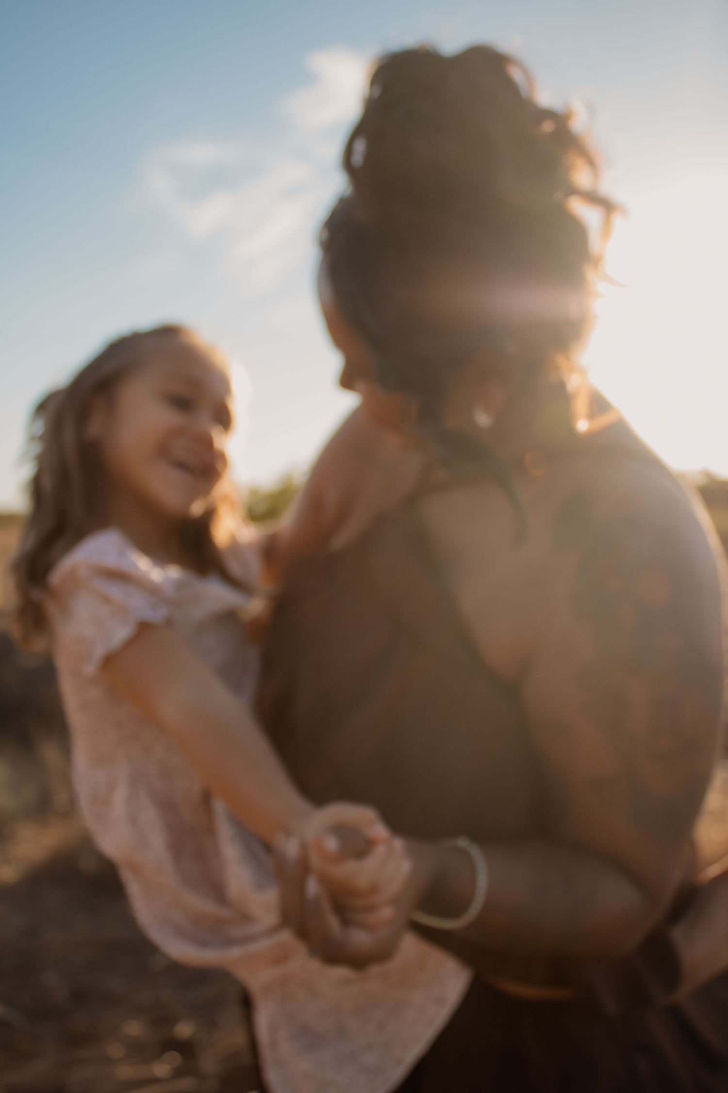 A joyful young girl in a white dress holds hands with a woman outdoors in golden sunlight.