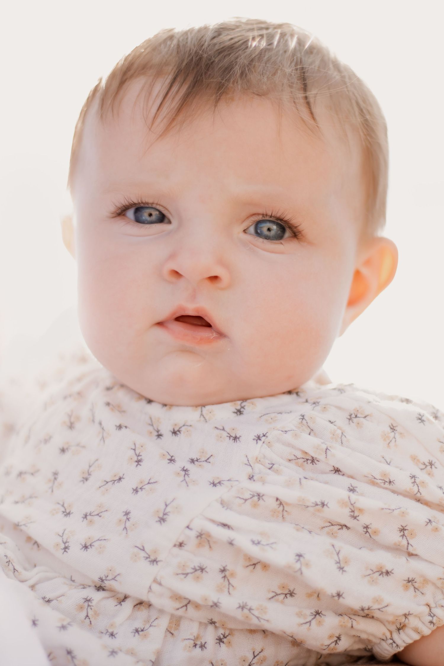 Adorable blue-eyed baby in a floral outfit gazing curiously at the camera against a white background.